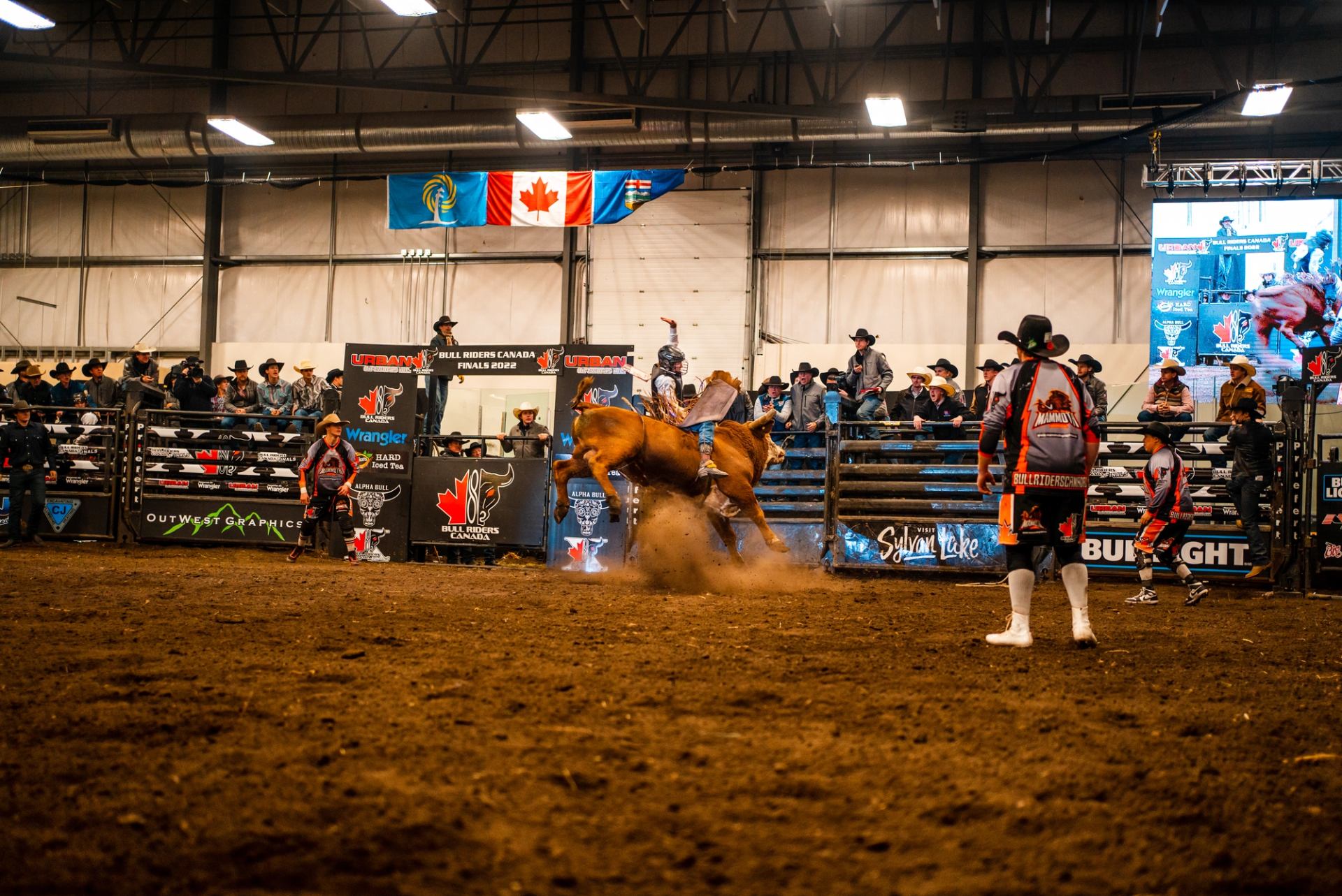 Indoor rodeo arena with bull rider, rodeo clowns, spectators and Canadian flags overhead