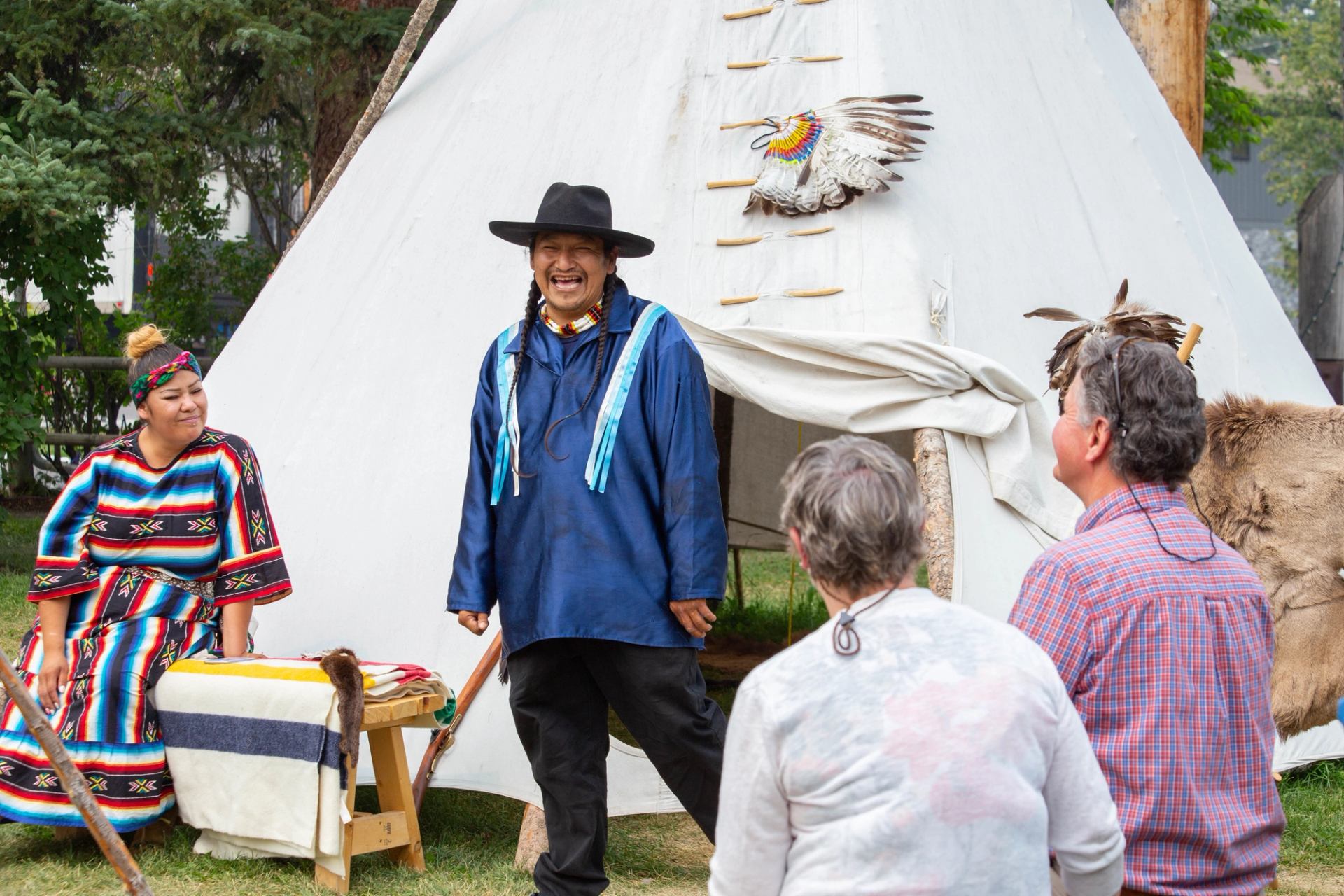 A smiling man in traditional attire stands outside a tipi, engaging visitors while another sits nearby in regalia.