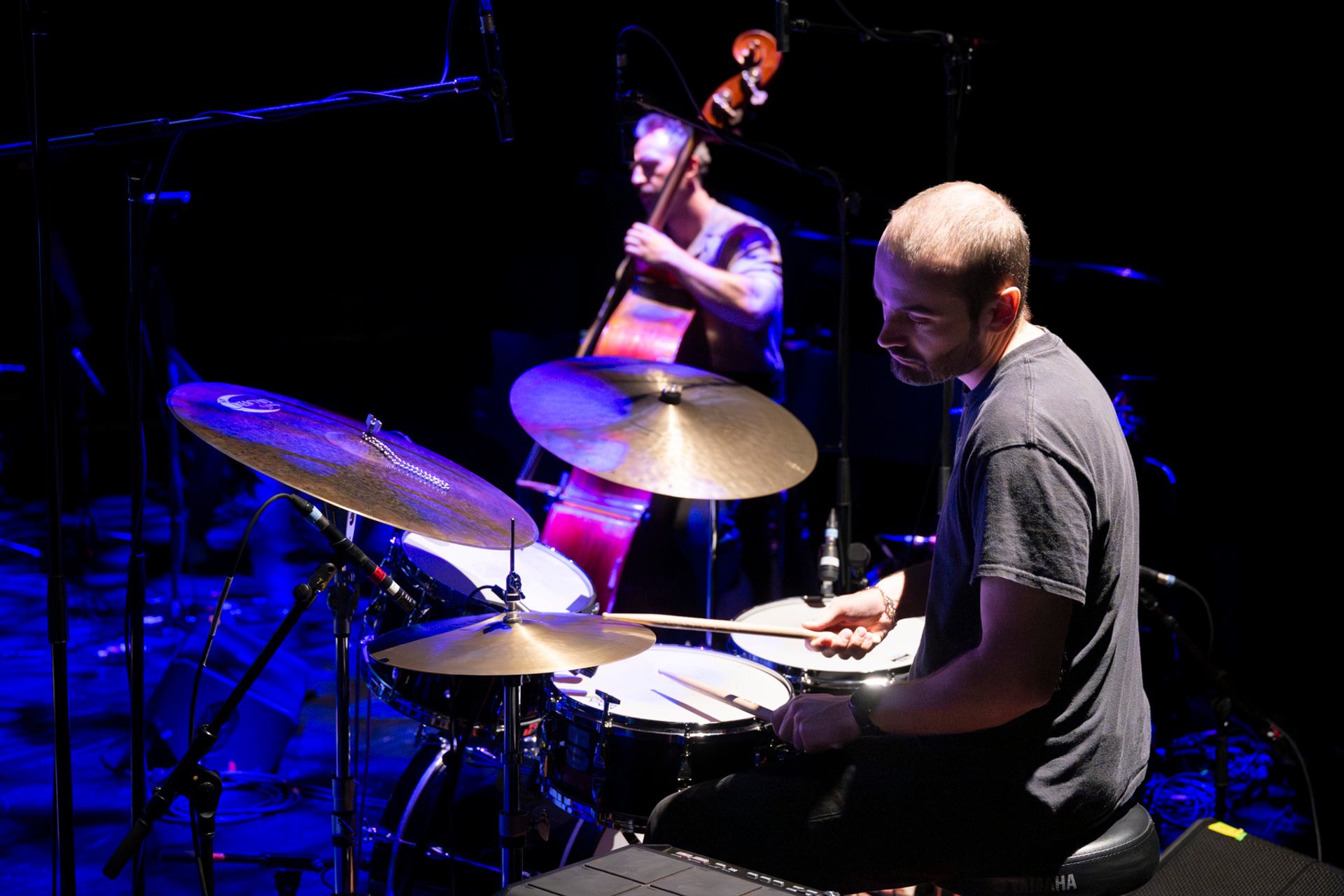 Drummer plays drum kit onstage while upright bassist performs in blue stage lighting.
