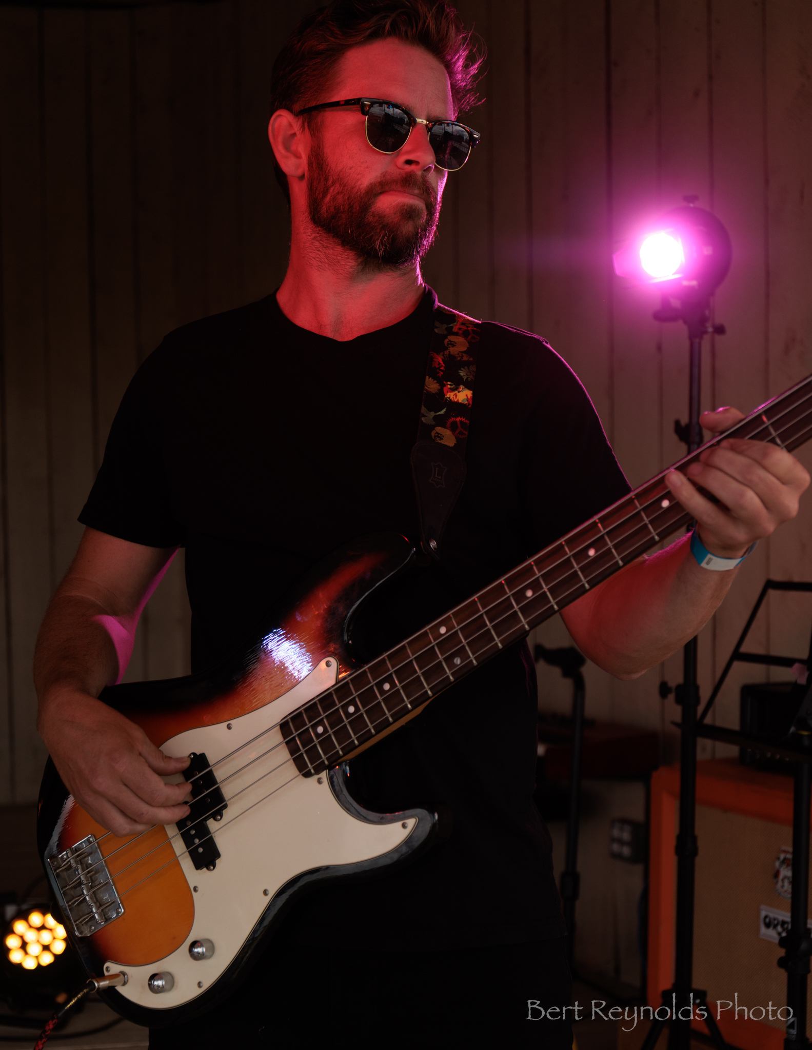 Musician wearing sunglasses plays electric bass guitar on stage under pink spotlight.