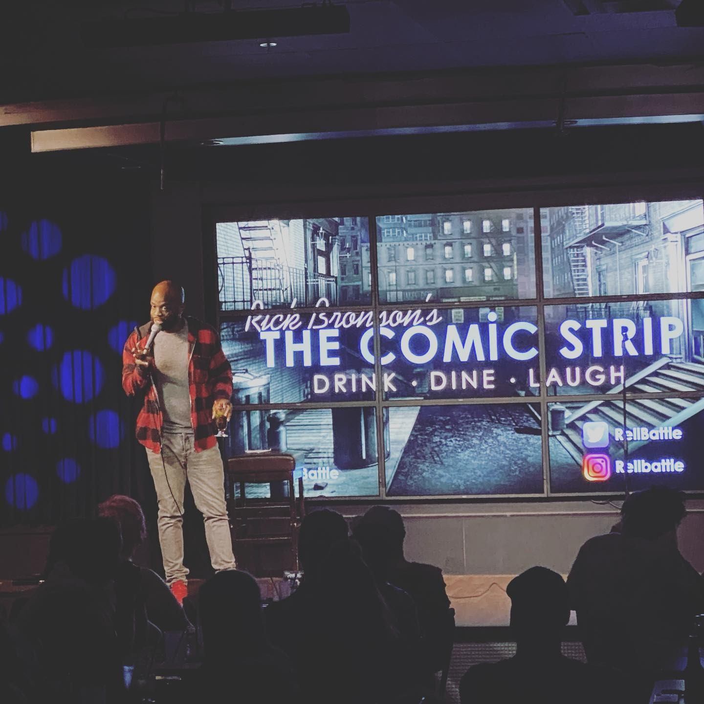 Comedian performing on stage beneath The Comic Strip signage with an engaged audience seated.