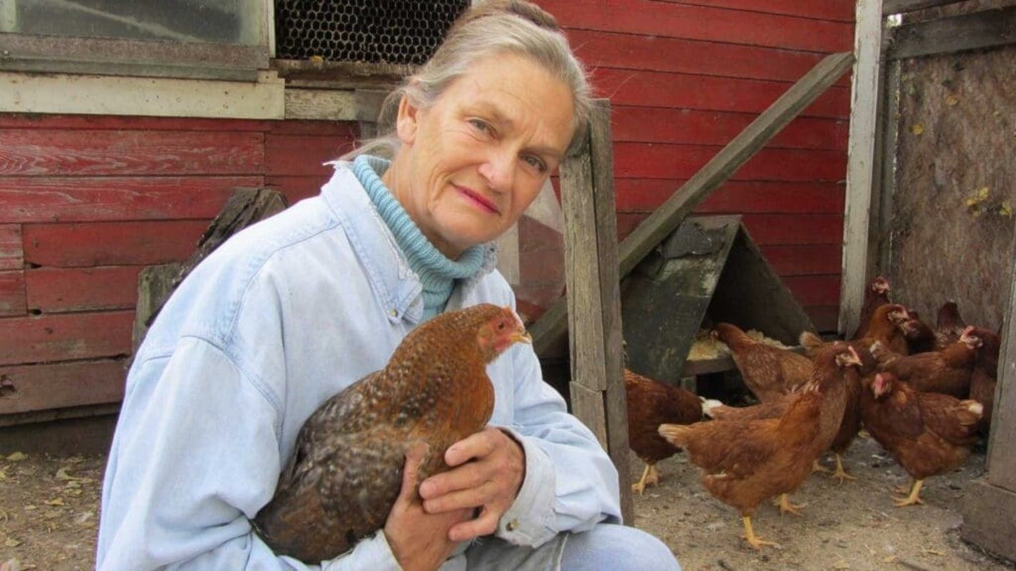 Farmer in denim shirt holding a brown chicken beside coop with flock in background.