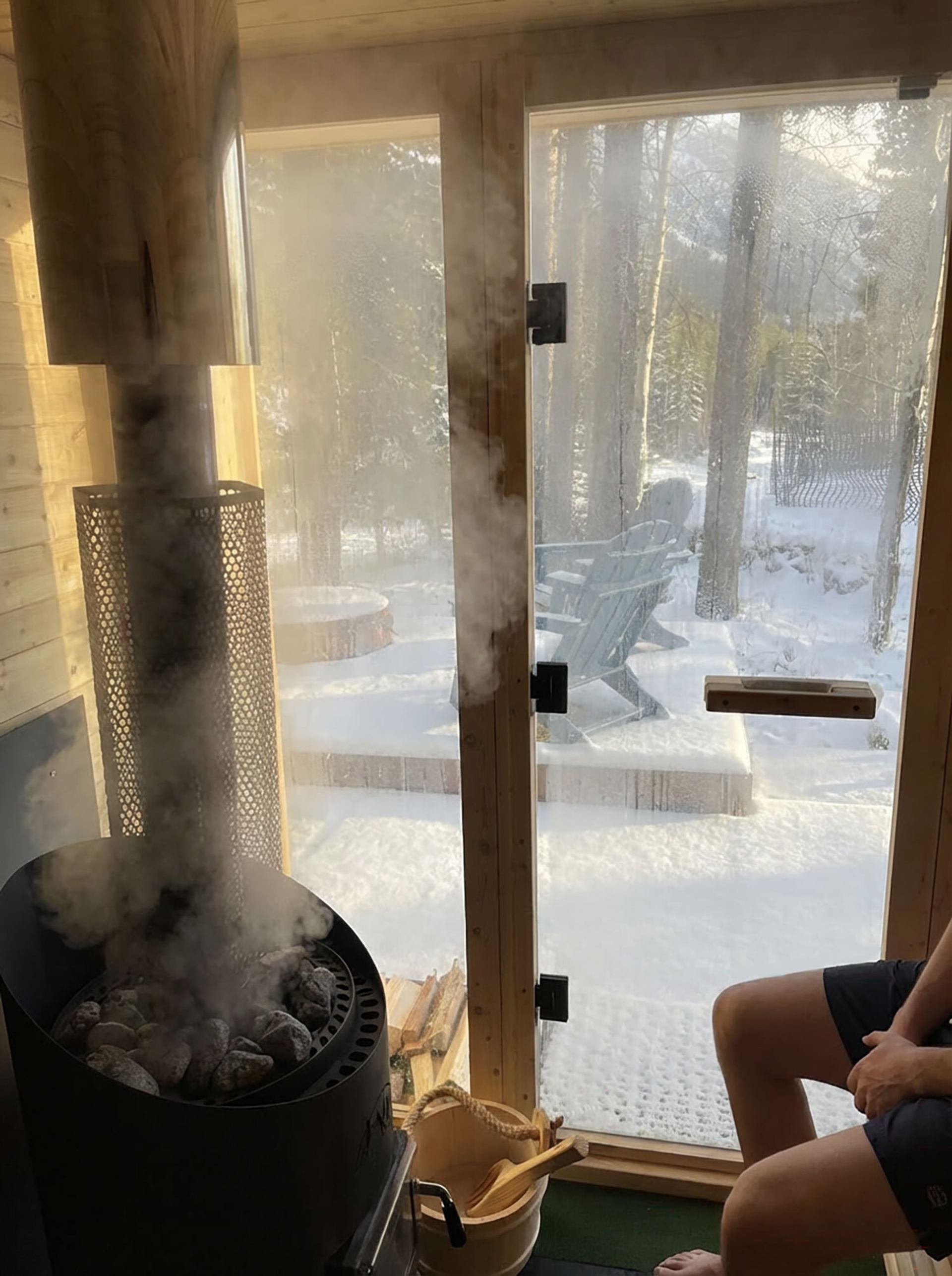 Sauna interior with steam rising, looking out to snow-covered trees and outdoor chairs.
