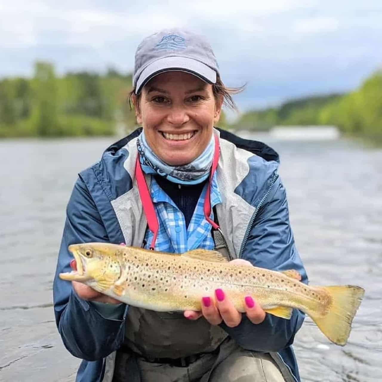 A smiling woman in a fishing cap and jacket holds a large brown trout.