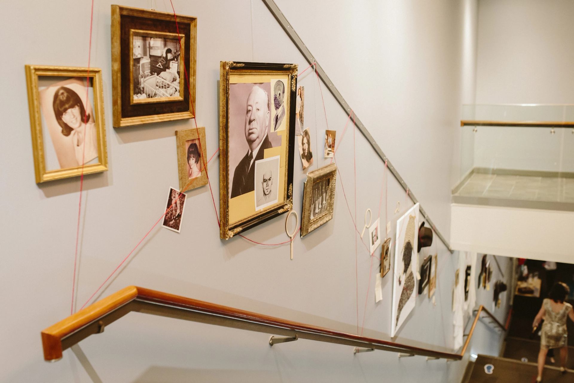 Stairwell wall display with framed portraits and photos connected by red string.