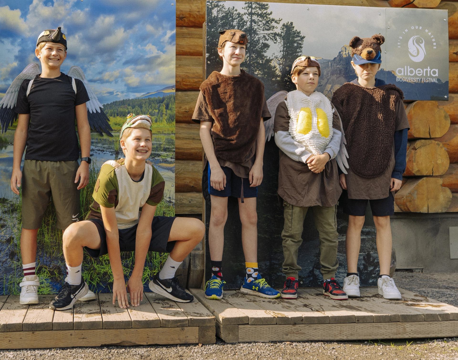 Five children wear animal-themed costumes and stand on a wooden platform in front of a nature backdrop.