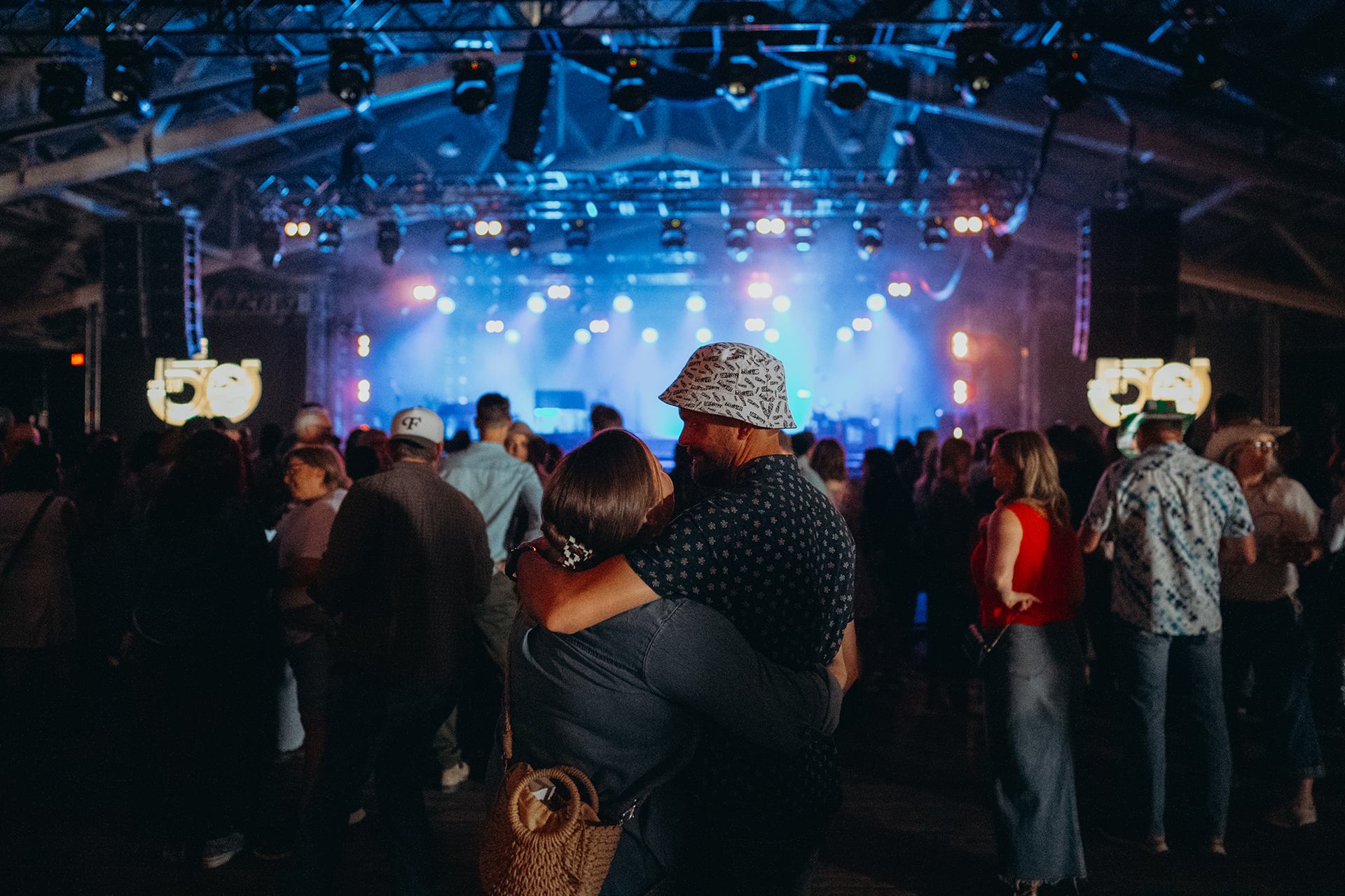 Crowd gathers in dimly lit concert hall, couple embracing near stage with blue lights.
