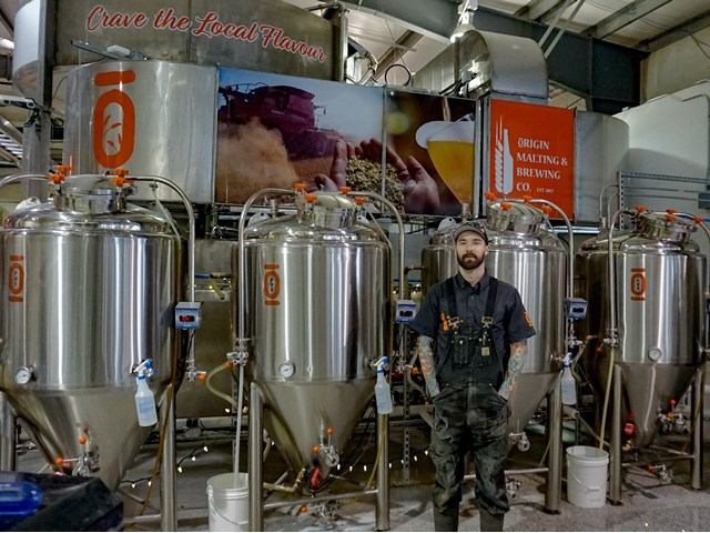 A man stands in front of a large brewery, showcasing its impressive structure and industrial atmosphere.
