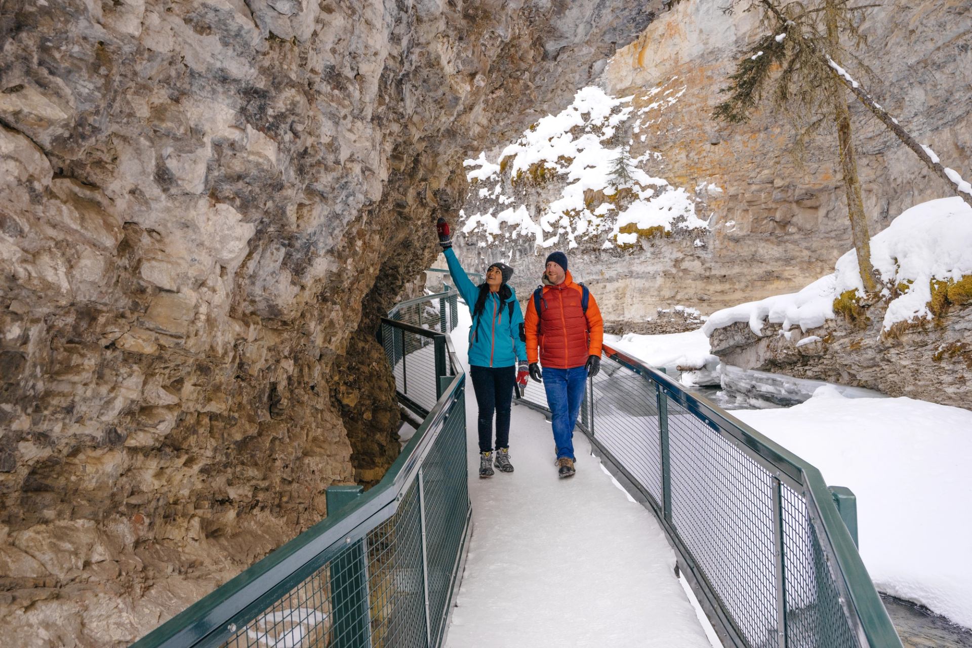 Two people walk along a narrow icy walkway beneath an overhanging canyon wall.