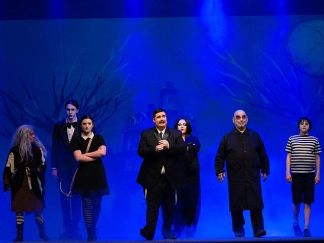 Actors walking on a stage under blue lighting with painted backdrop trees and figures.