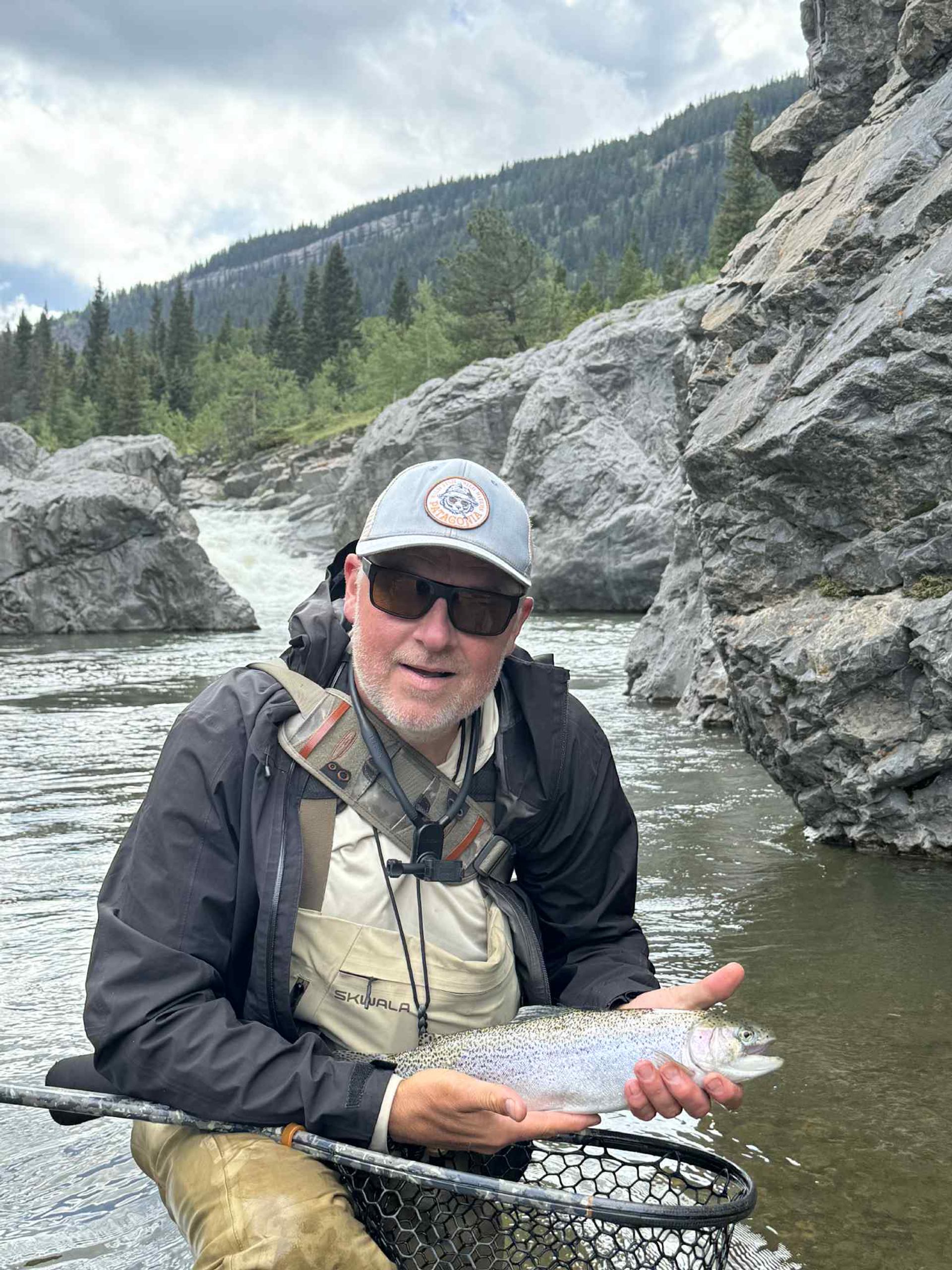Man in waders holding a rainbow trout in a rocky river with forested mountains.