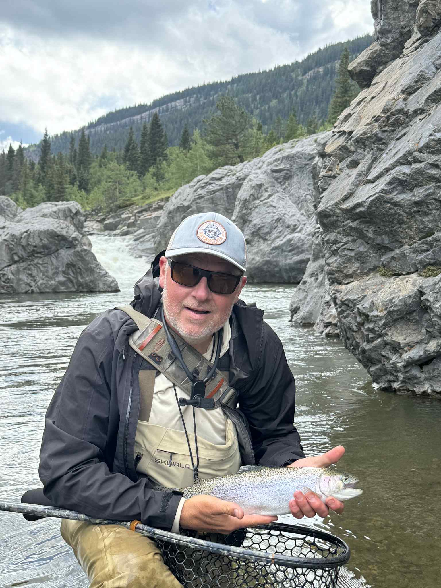Man in waders holding a rainbow trout in a rocky river with forested mountains.