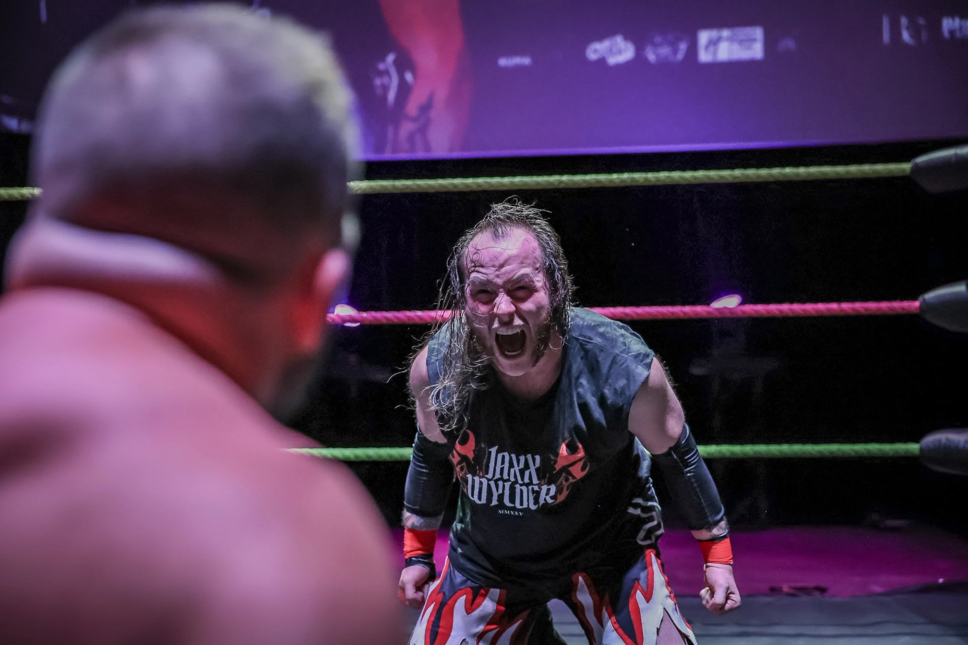 Professional wrestler in flame-patterned tights yells at opponent inside brightly lit ring.