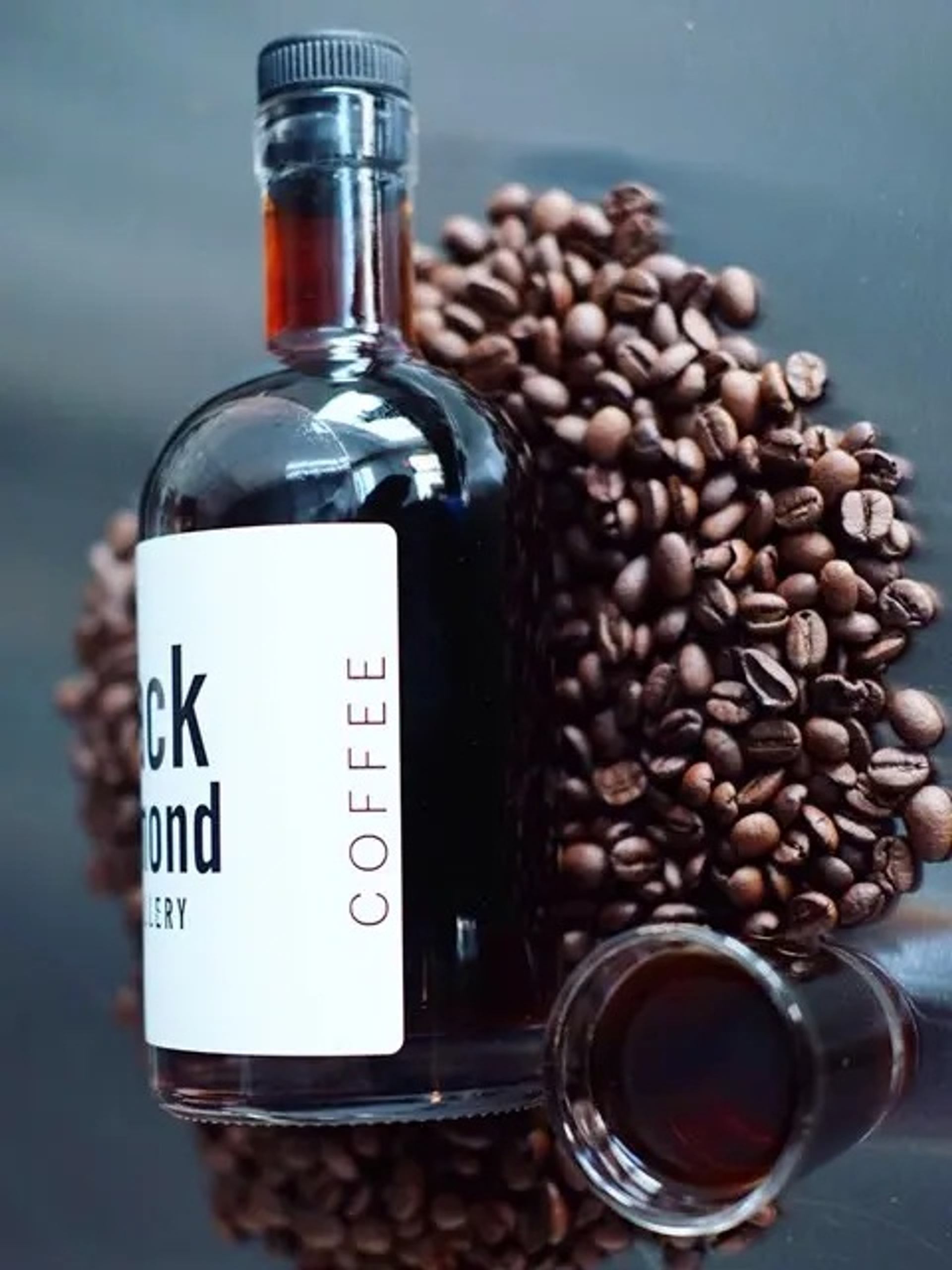 Glass bottle of coffee liqueur surrounded by roasted coffee beans and a filled shot glass.