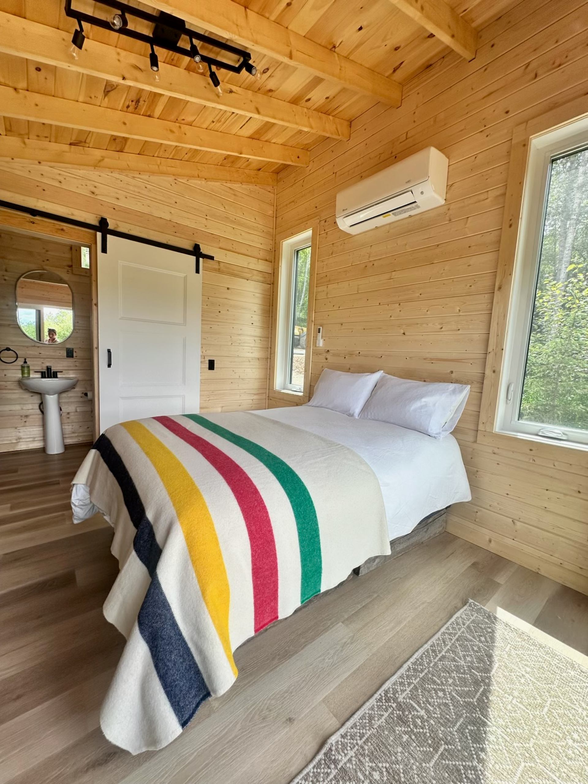 Bright cabin bedroom with wood walls, a bed with a striped blanket, and a door to a small bathroom.