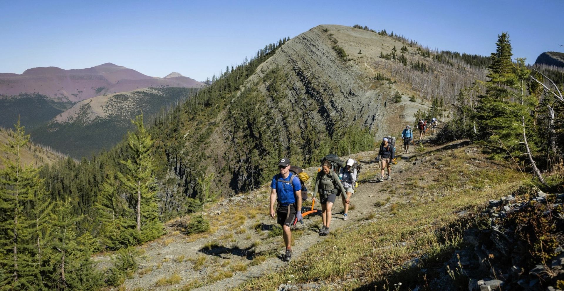 Hikers with backpacks walk along a narrow mountain ridge trail above forested valleys.