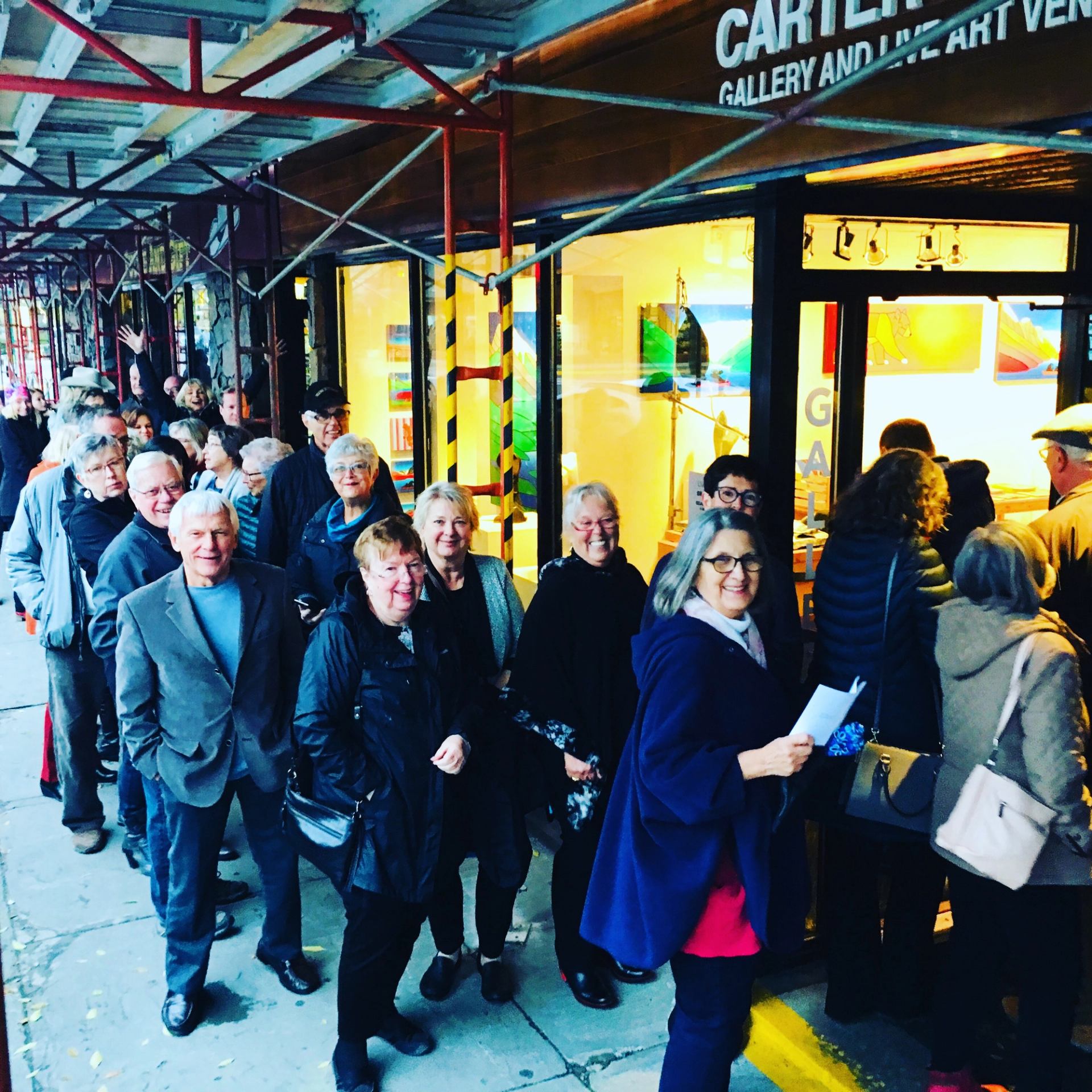 People lined up outside brightly lit gallery under scaffolding.
