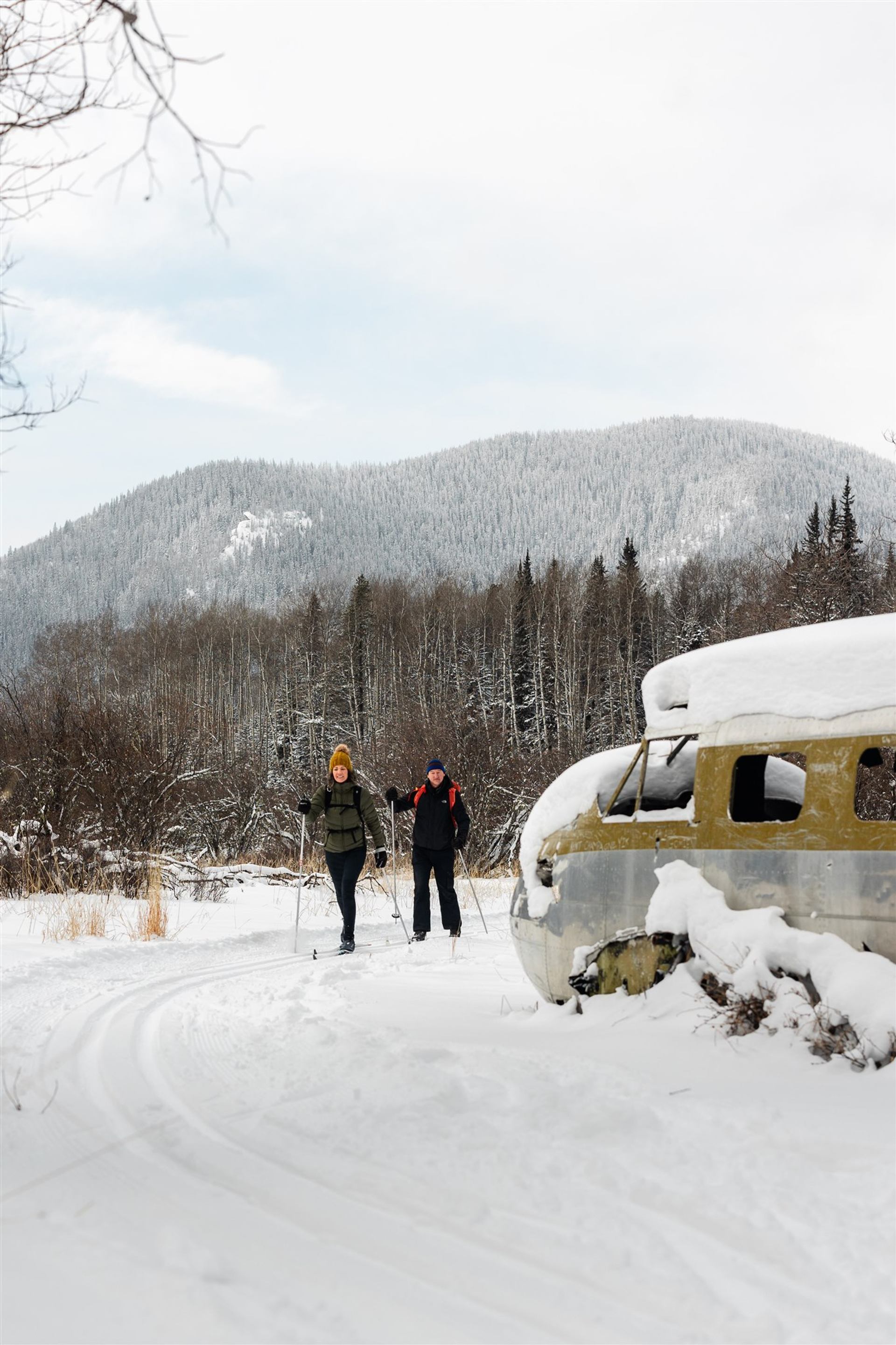 Two skiers pass a snow-covered old vehicle in a winter valley with forest and mountains.