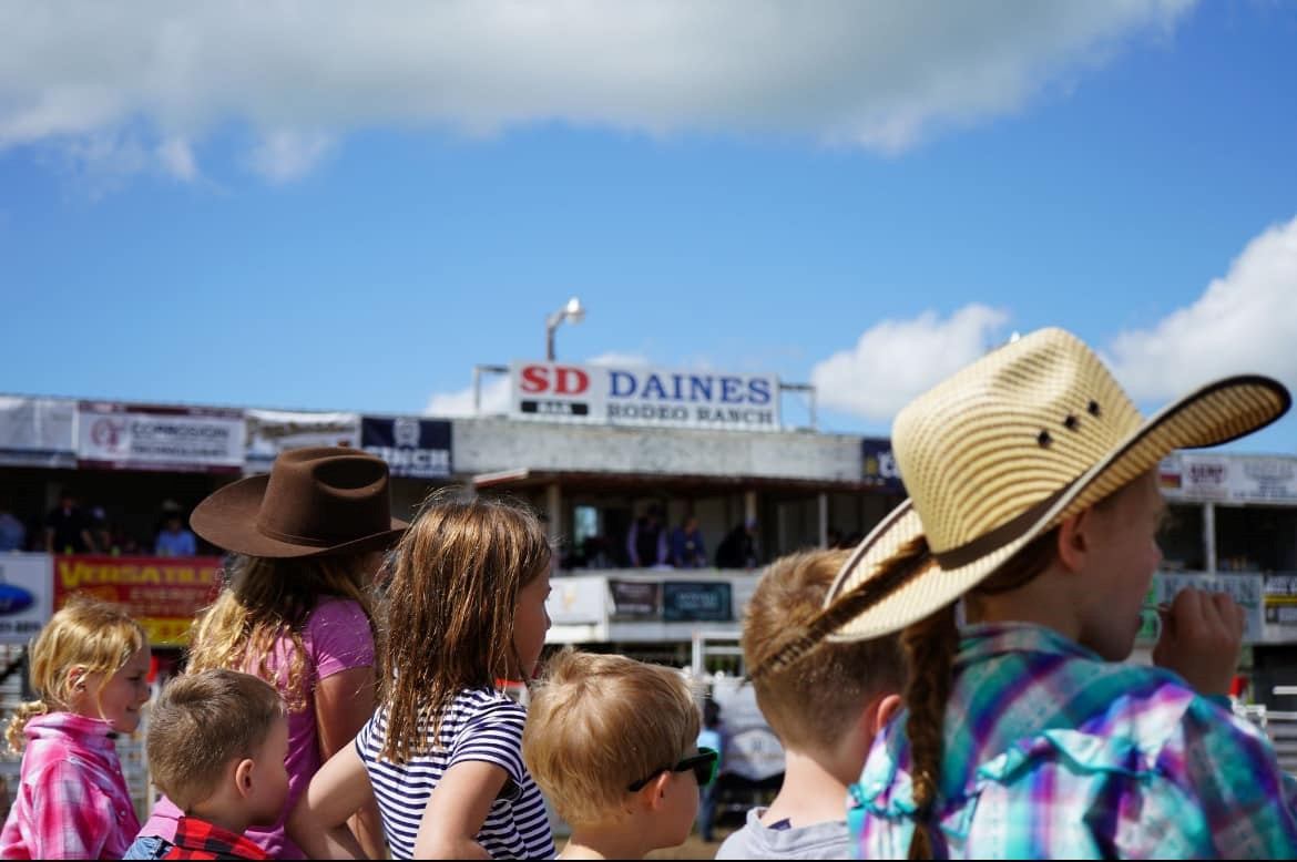Children watching the rodeo from the rail with the Daines Ranch Rodeo Ranch grandstand and signage in the background.