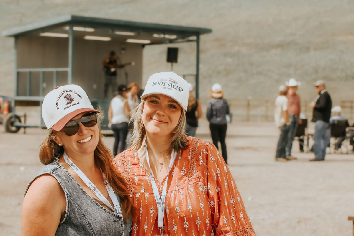 Two women smiling in ball caps on a sunny day outside. There is a crowd and a guitar player behind