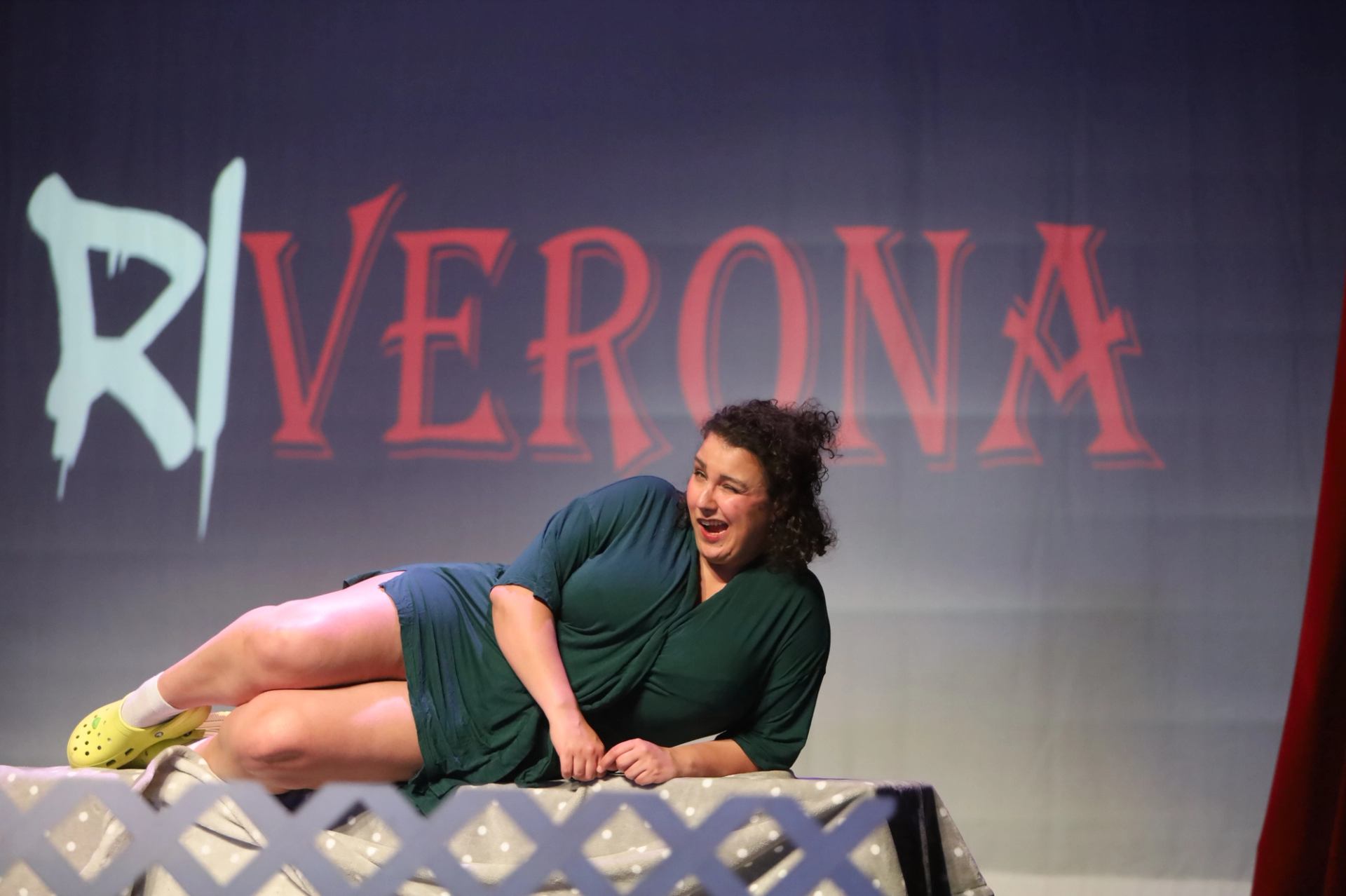 Stage performance with a person reclining in front of a backdrop reading “RIVERONA.”