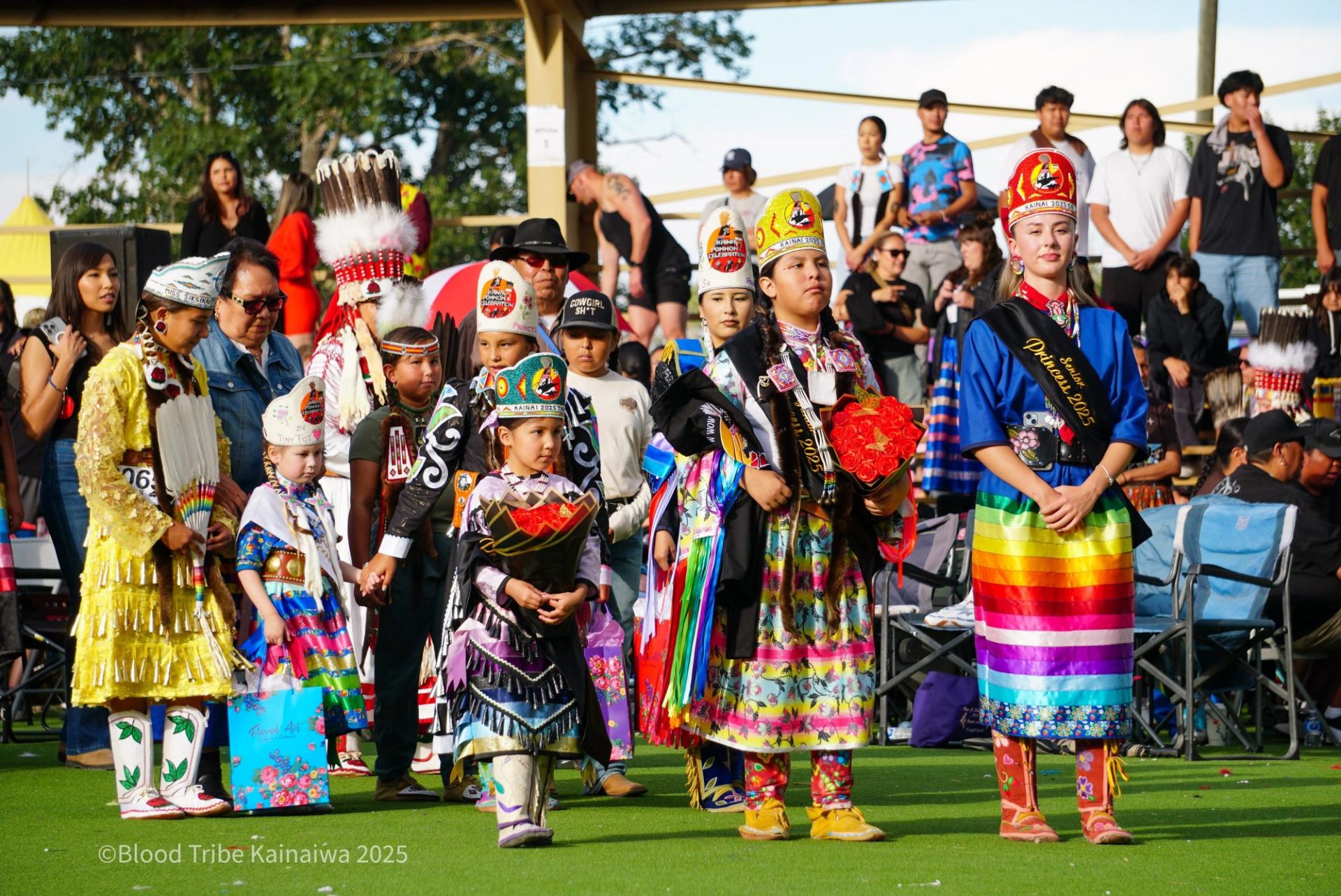 Children in colourful regalia stand on grass during a crowded outdoor gathering.