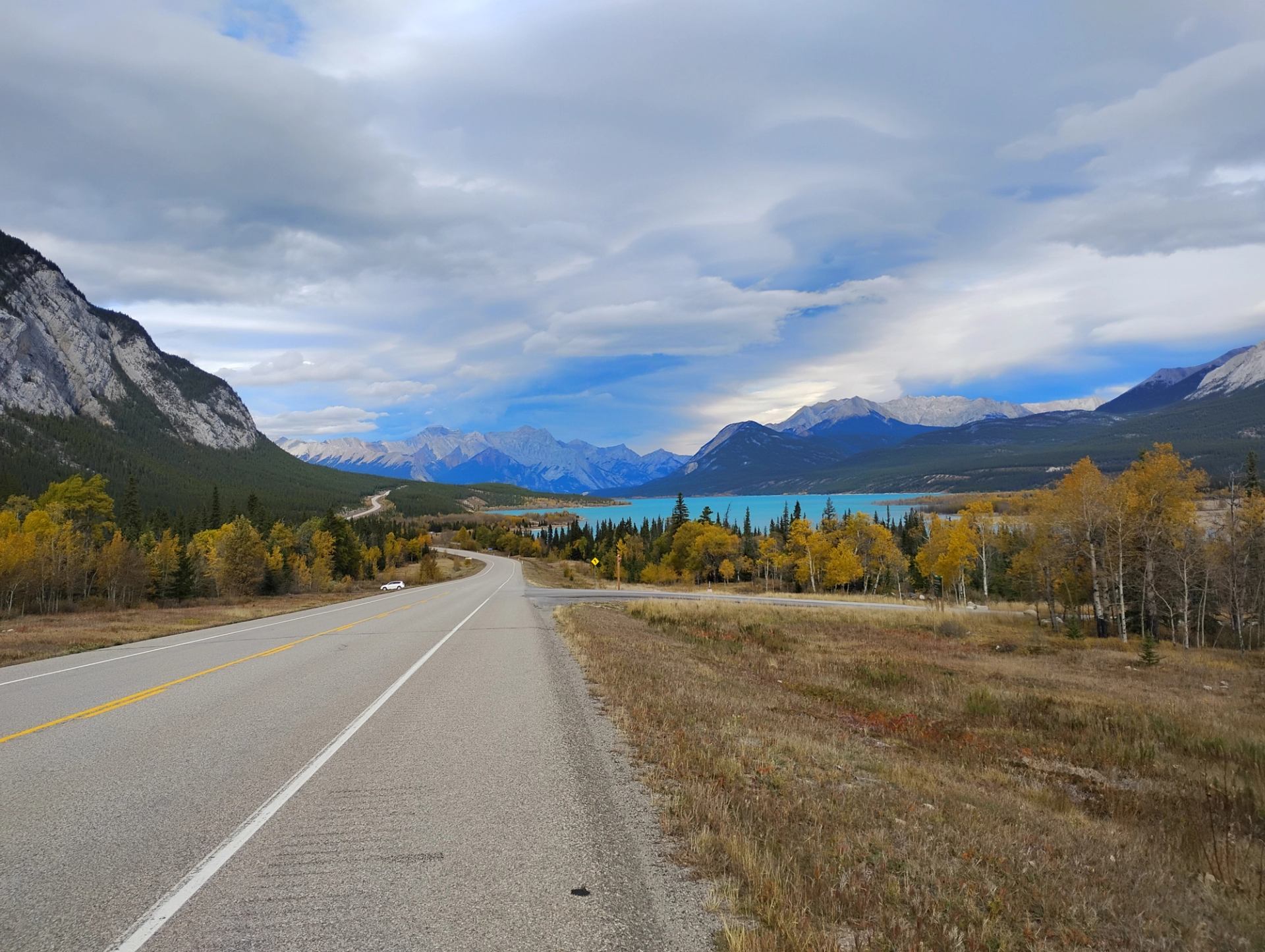 Open highway overlooking a turquoise lake and mountain ranges under a cloudy sky.