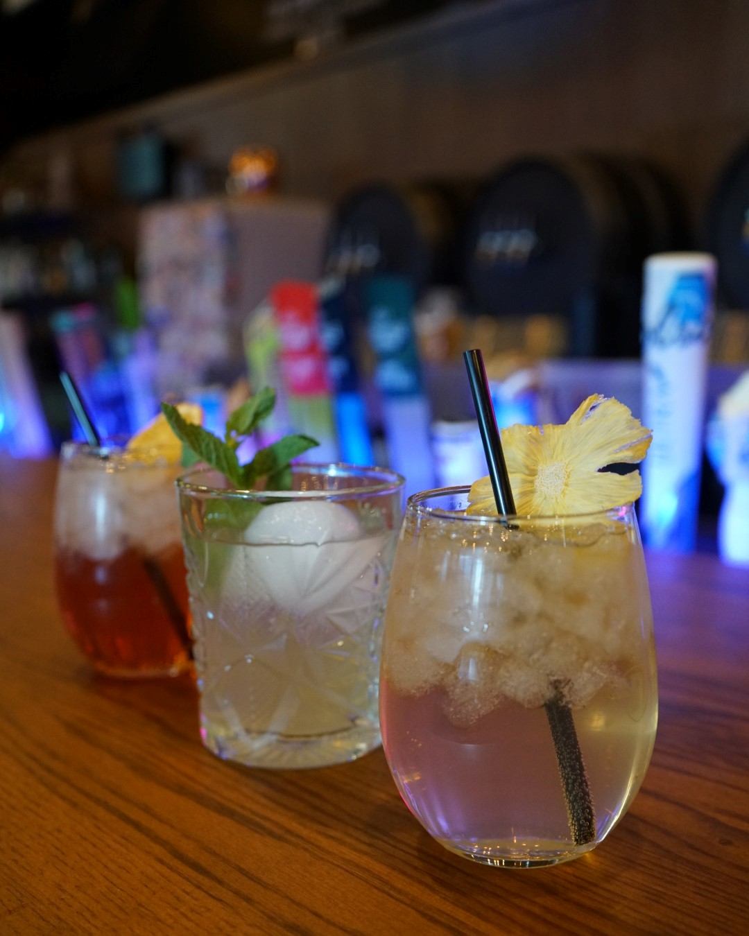 Cocktails with straws and pineapple garnish on a wooden bar, bottles blurred behind.