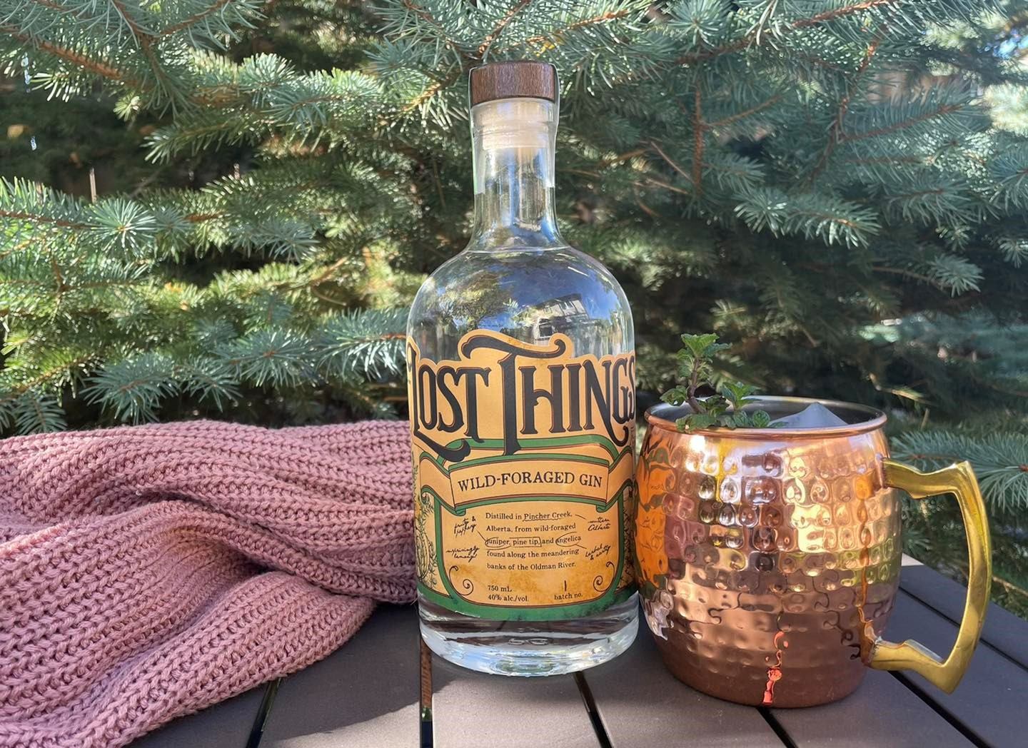 A bottle of Lost Things gin next to a copper mug on a picnic table in front of a pine tree.