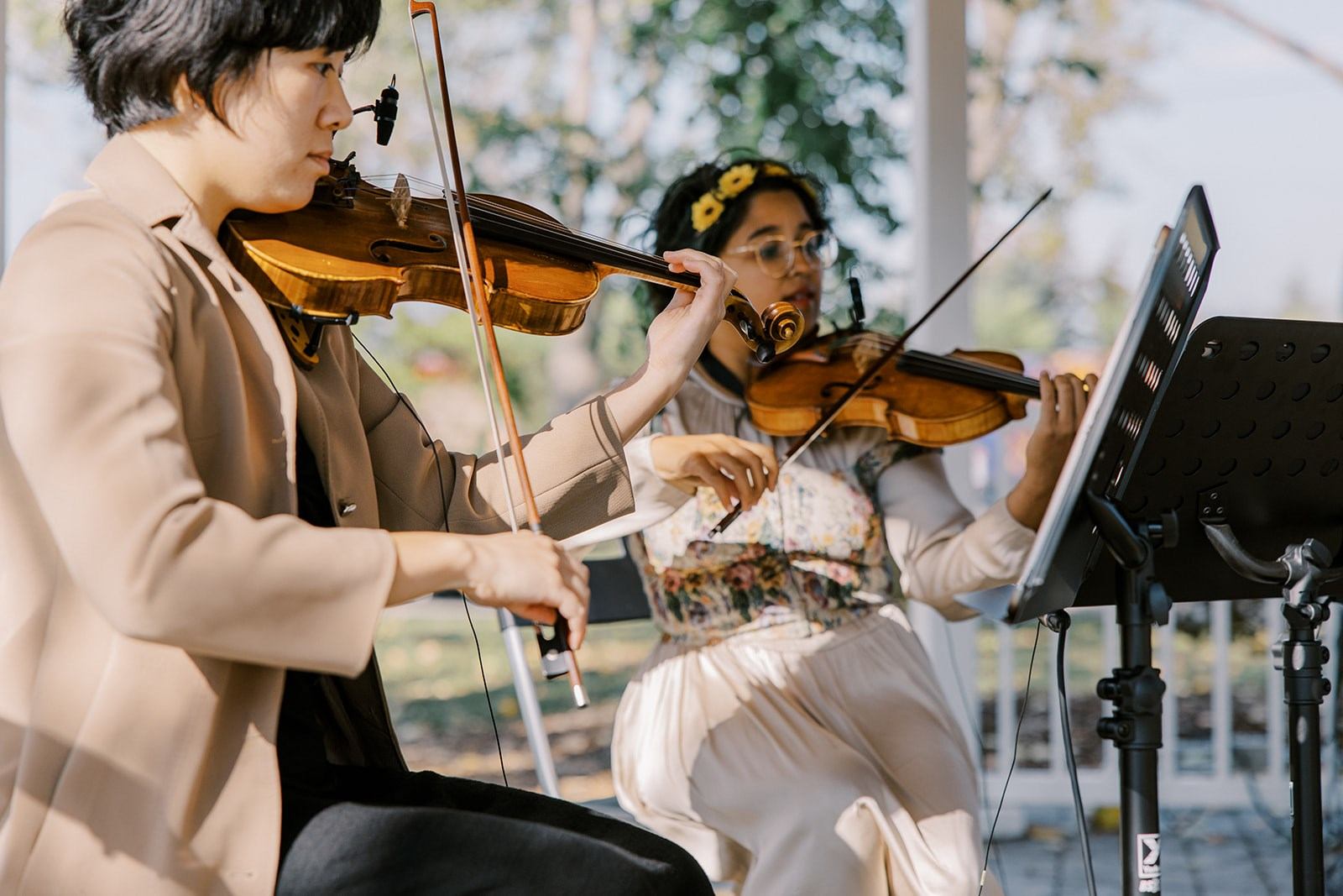 Two violinists play beside a music stand in an outdoor covered area.