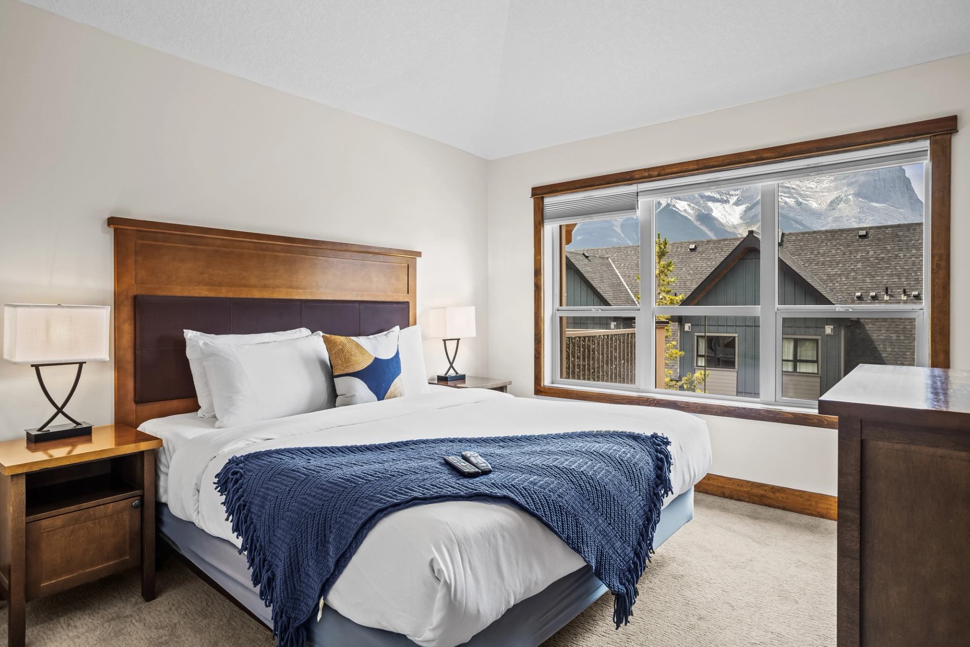 Bedroom with white bedding, blue throw, wooden furniture and mountain view through window.