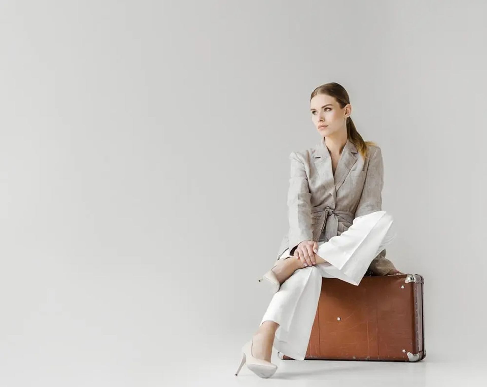 Person in a grey blazer and white trousers sits on a brown suitcase.