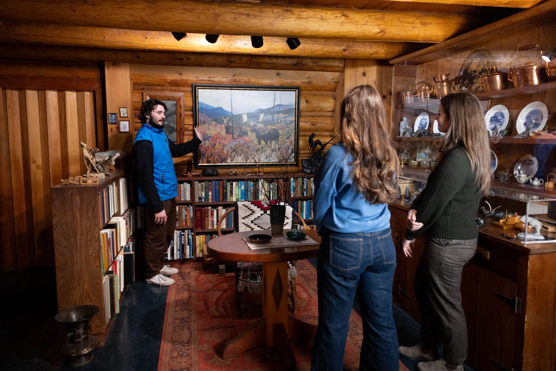 A guide gestures to framed artwork while visitors listen inside a log cabin room.