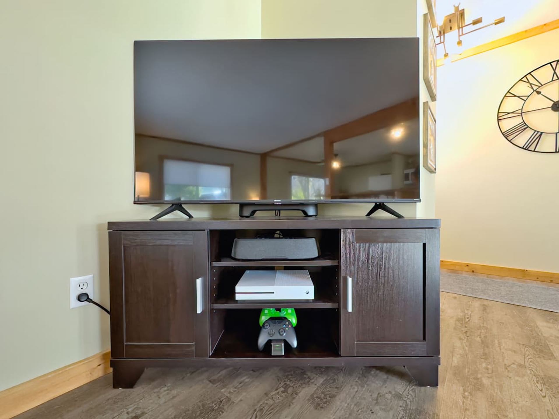 A large TV sits on a dark brown entertainment stand holding a soundbar, a white Xbox One, and two Xbox controllers.