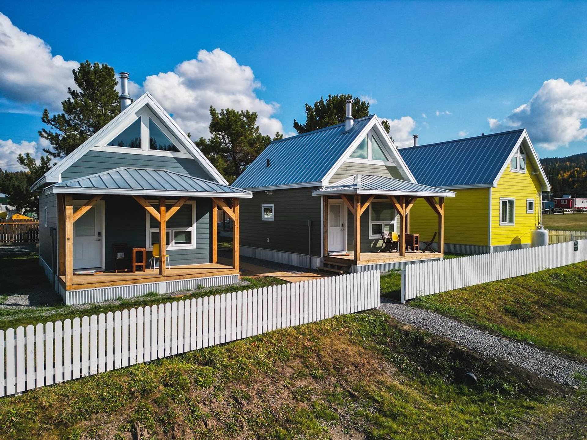Three small cottages with porches and a white picket fence on a sunny day.