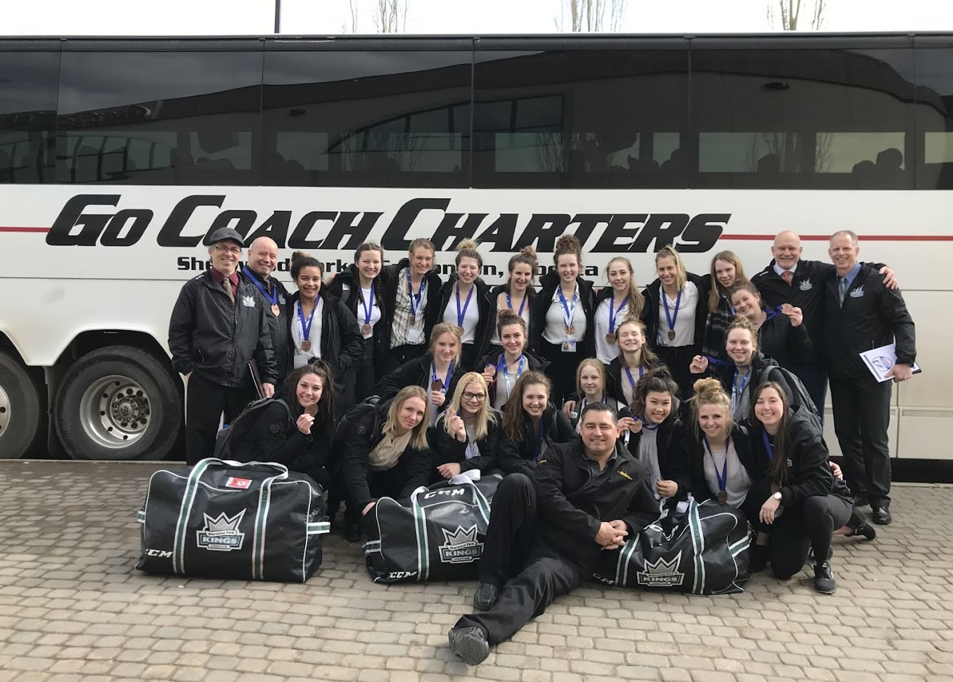 A group of athletes and supporters poses in front of a motorcoach with sports bags arranged in the foreground.