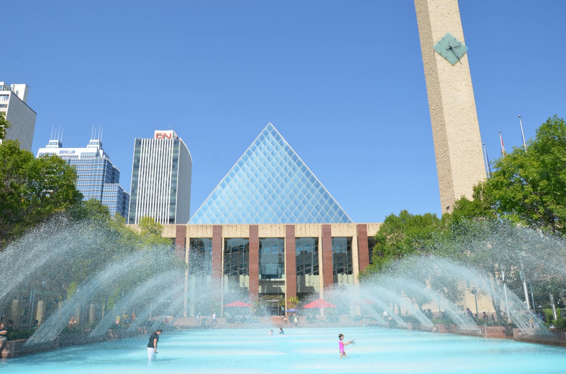 People playing in a large fountain in front of a building with a glass pyramid and a tall clock tower.