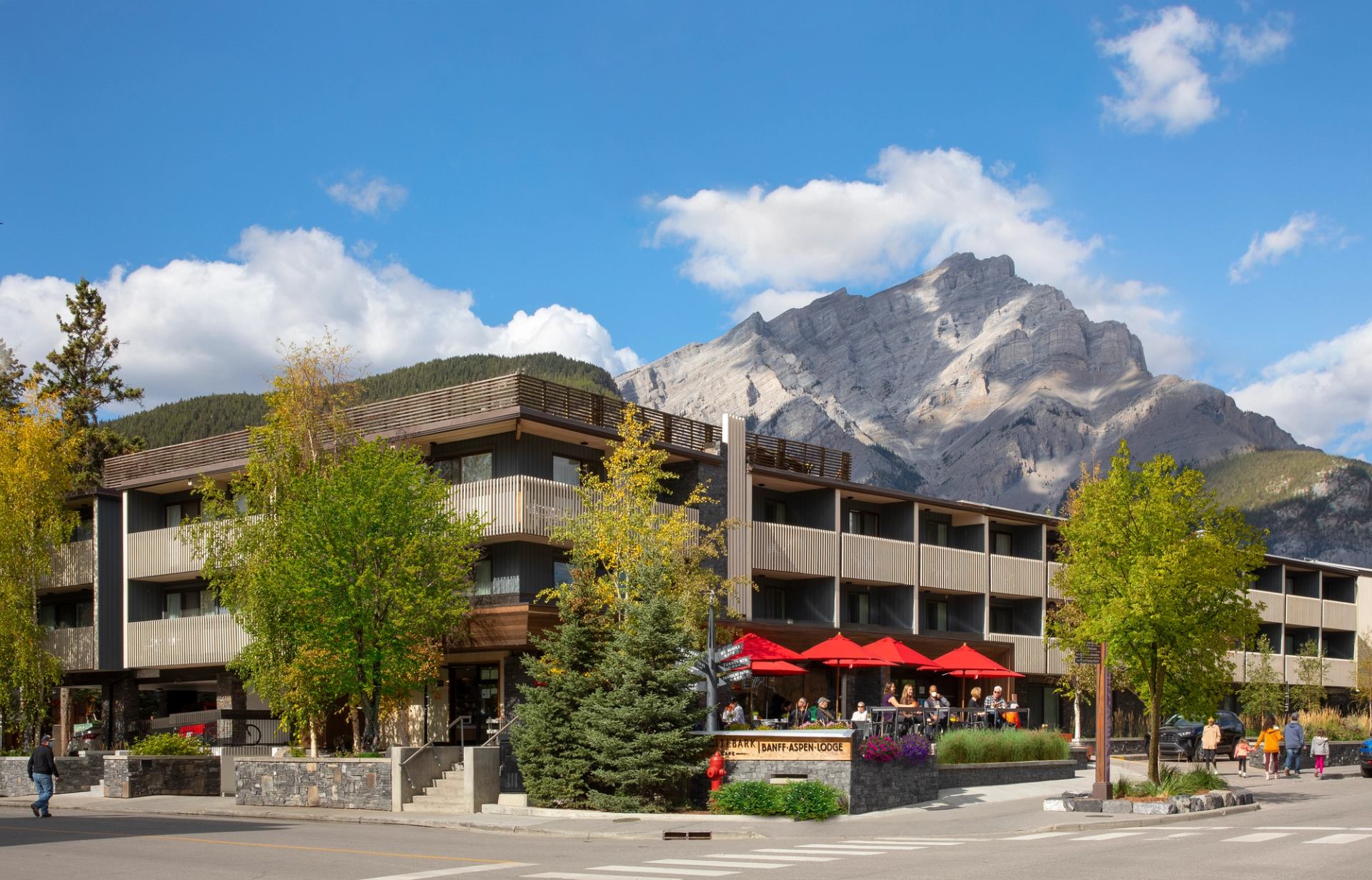 Modern hotel building with red patio umbrellas and towering rocky mountains behind