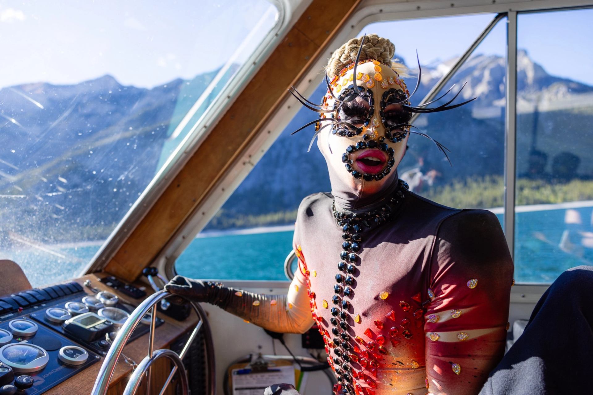 Person in ornate costume steers boat on lake, with mountains and water in the background.