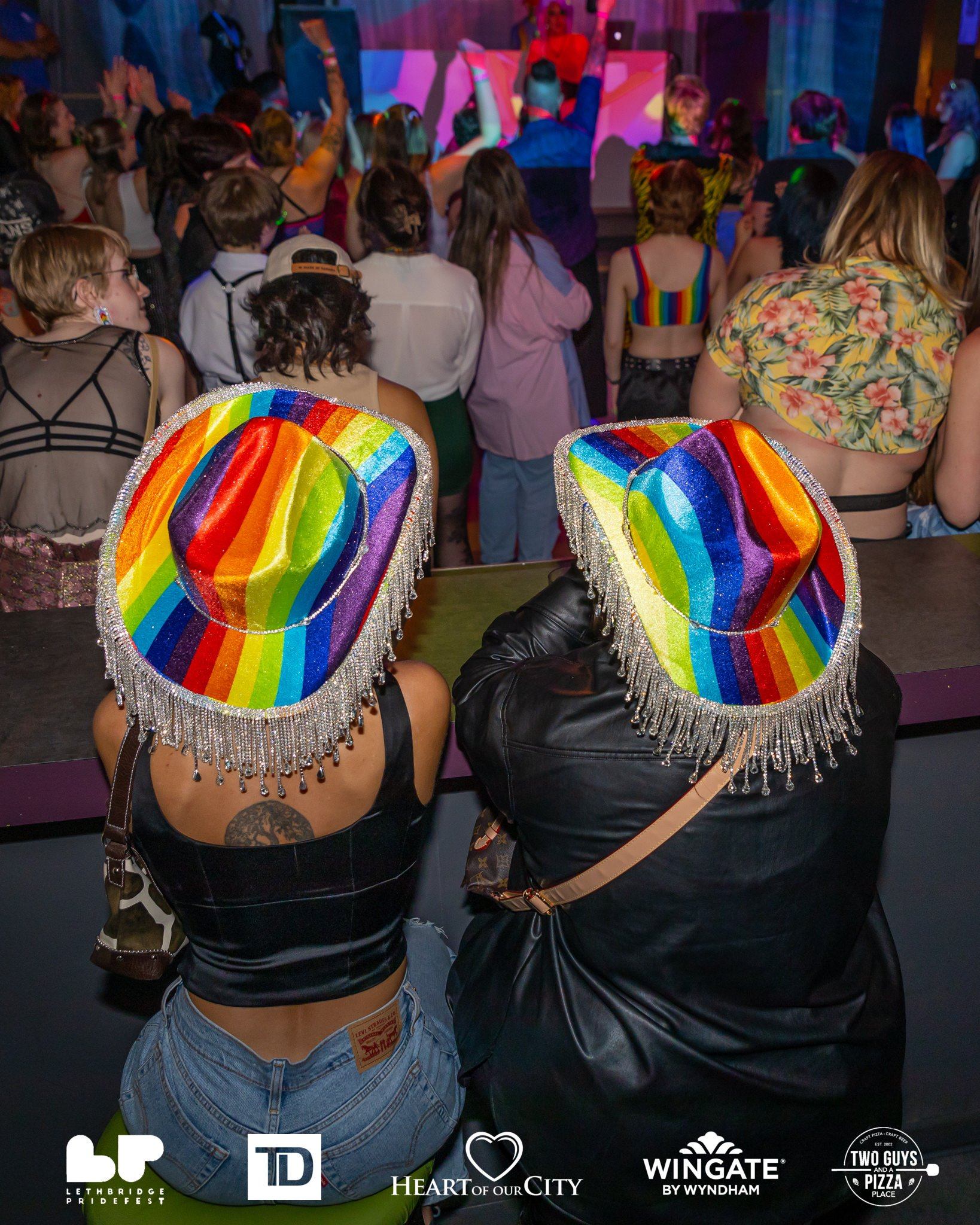 Two people wearing rainbow hats face a lively crowd dancing at Lethbridge Pride Fest.
