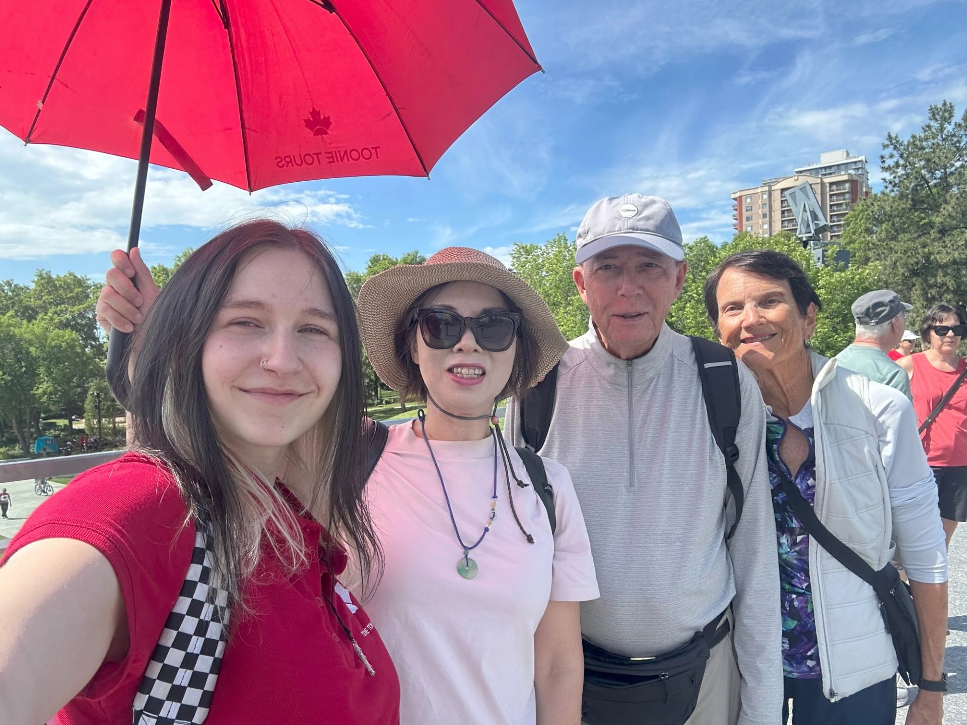 Four people outdoors with a red maple leaf umbrella under a blue sky in Edmonton.