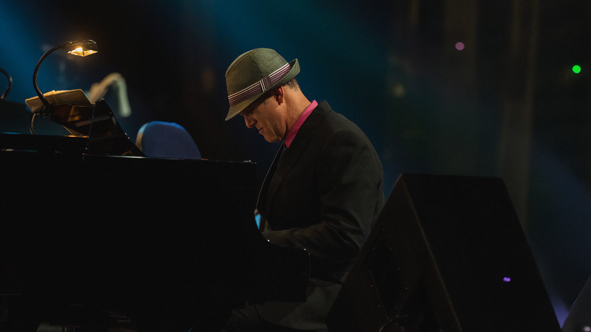 Pianist in suit and hat plays grand piano under stage light in dark venue.