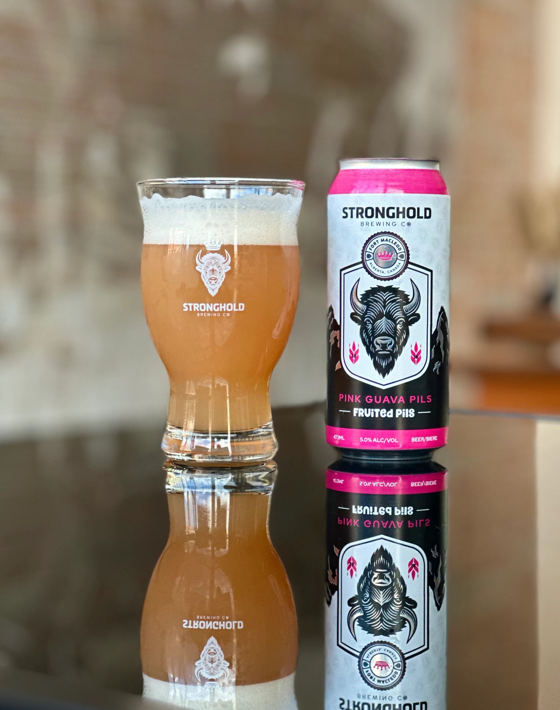 Glass of hazy pink pilsner beside a can of Stronghold Pink Guava Pils on a reflective tabletop.
