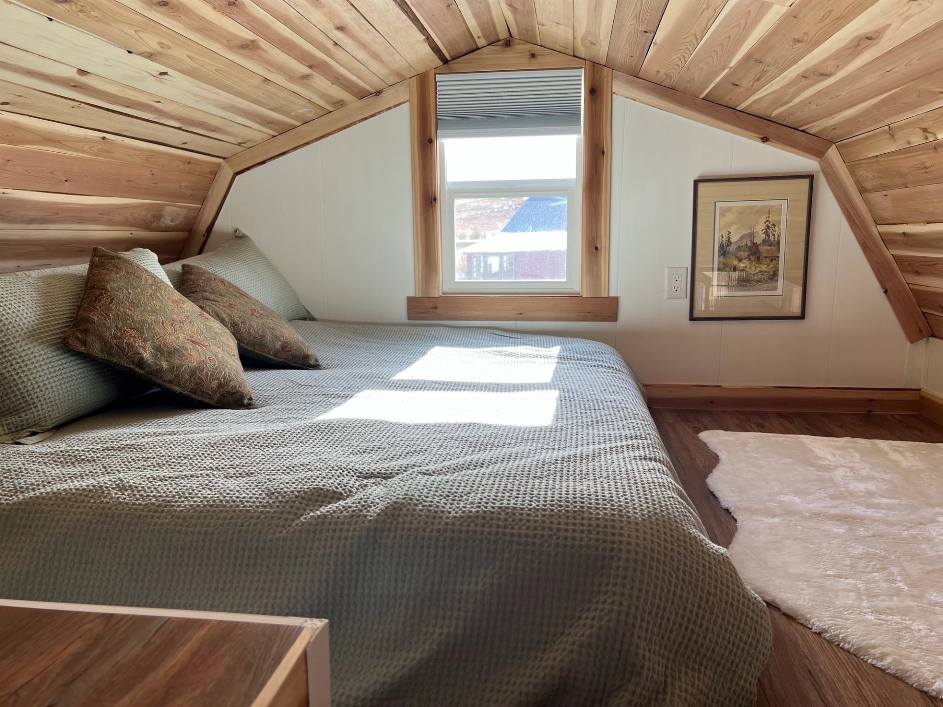 Loft bedroom with wood-paneled ceiling, a large bed, pillows, and a window letting in sunlight.