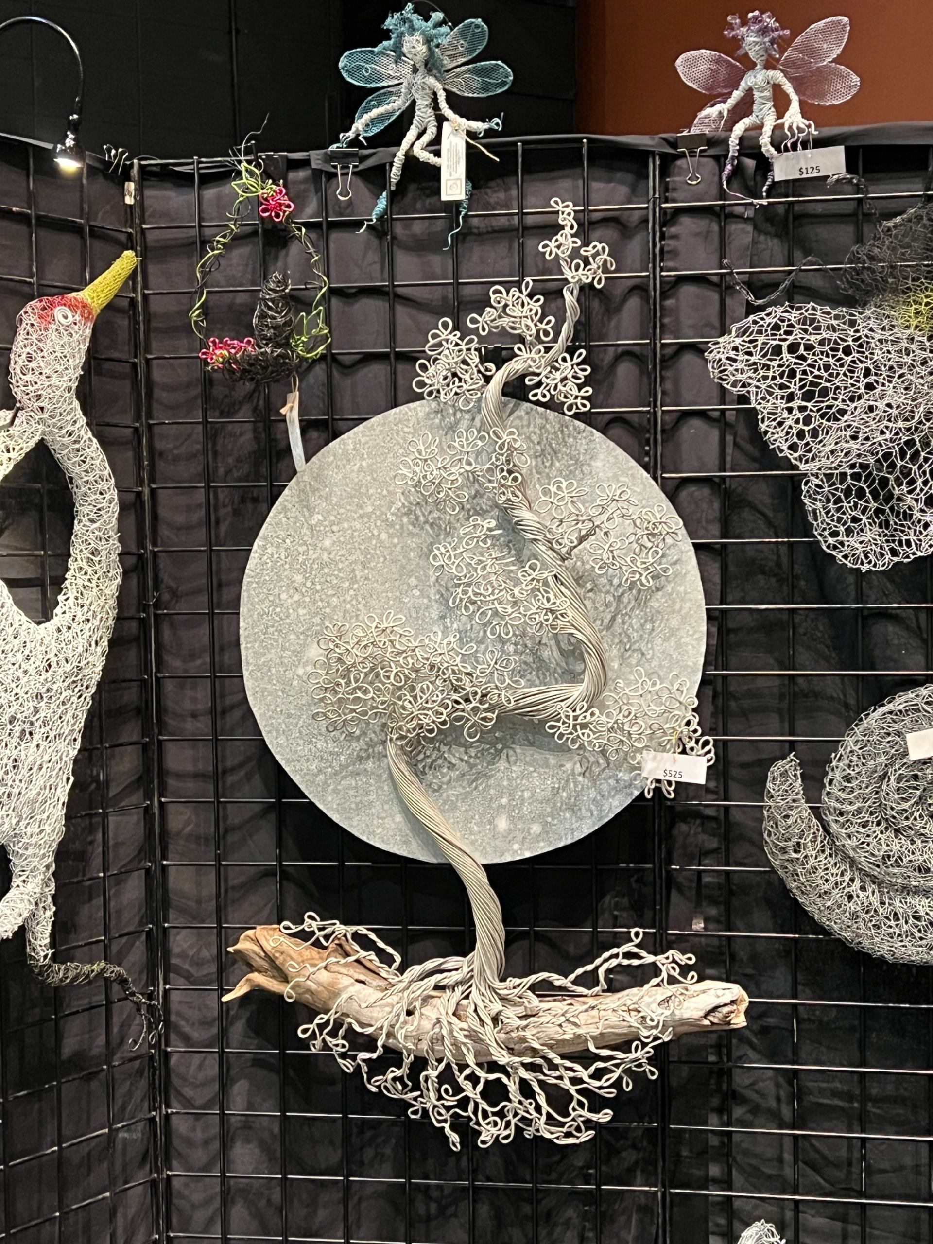 Wire tree sculpture on round metal disc, displayed on grid wall with price tags.