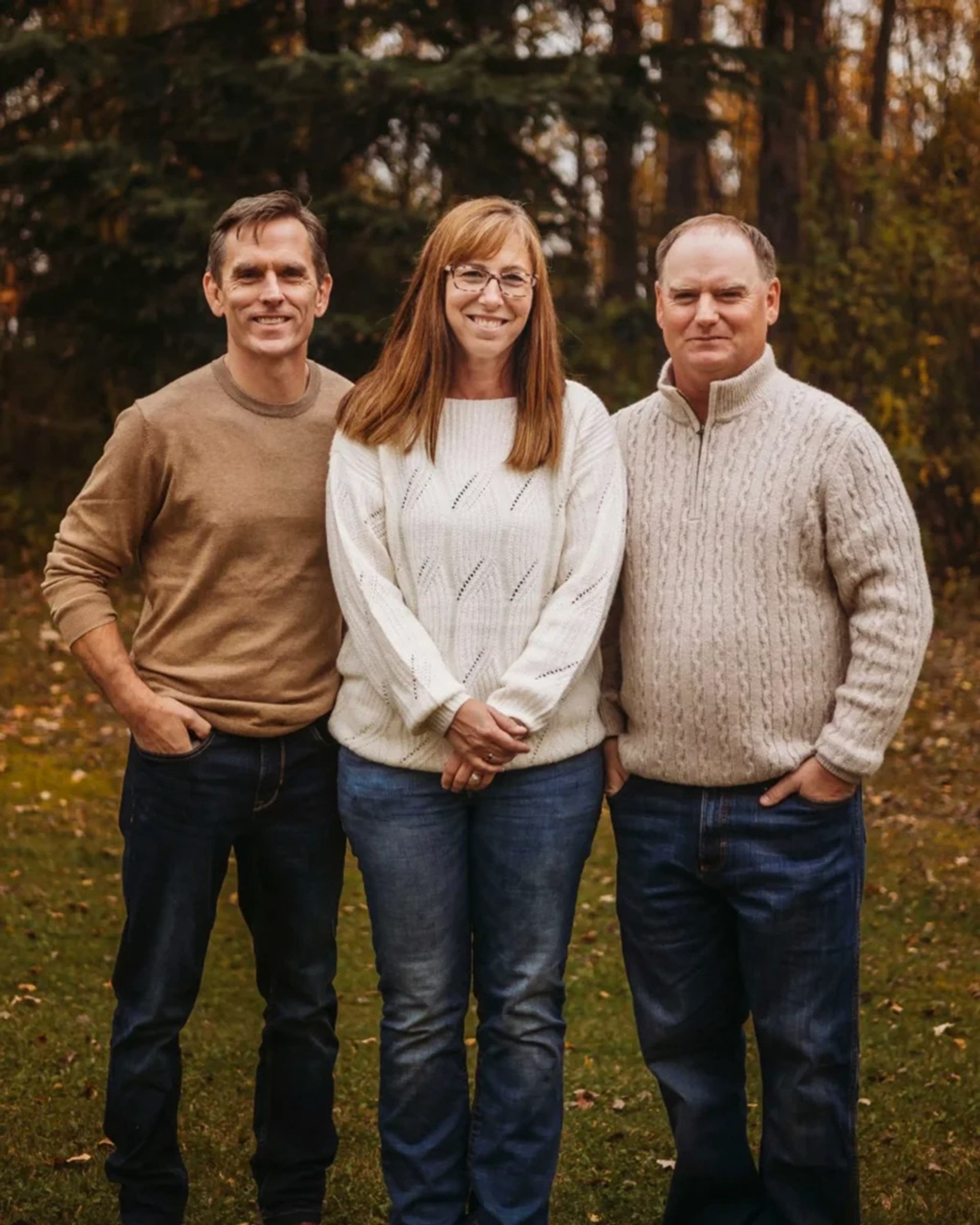 Three adults in sweaters stand on grass with trees in the background.