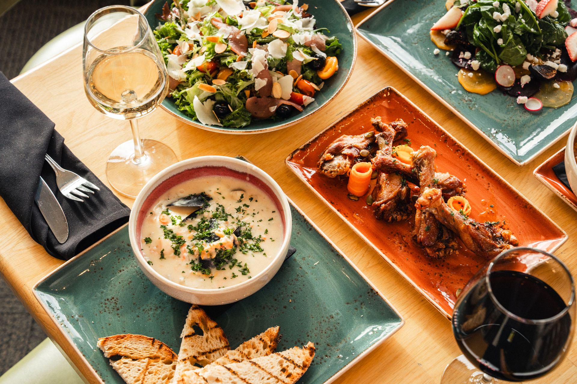 Restaurant table with salad, creamy soup, grilled bread, chicken wings and wine glasses.