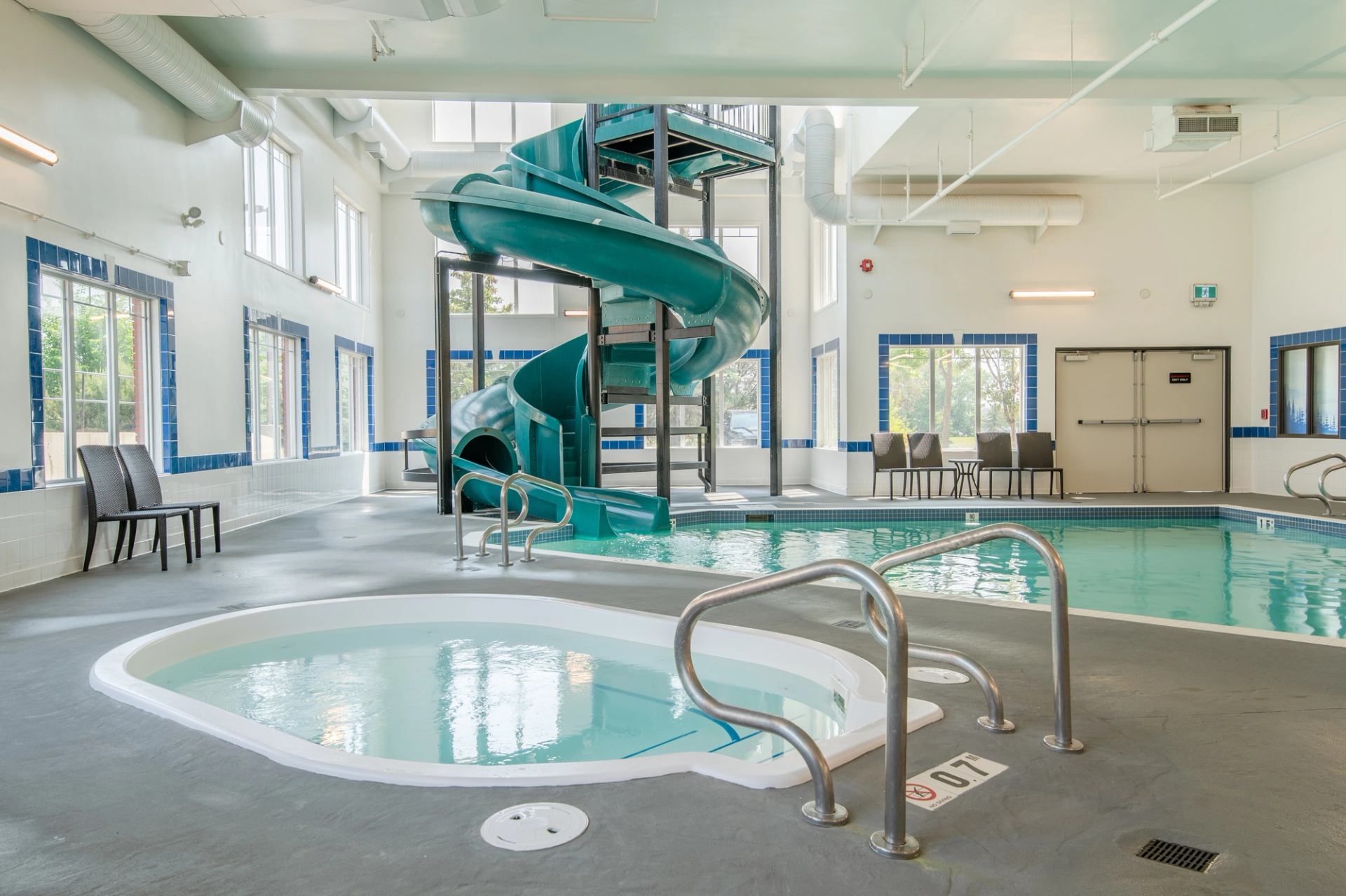Indoor pool area with waterslide, hot tub, pool ladders and chairs along windows.