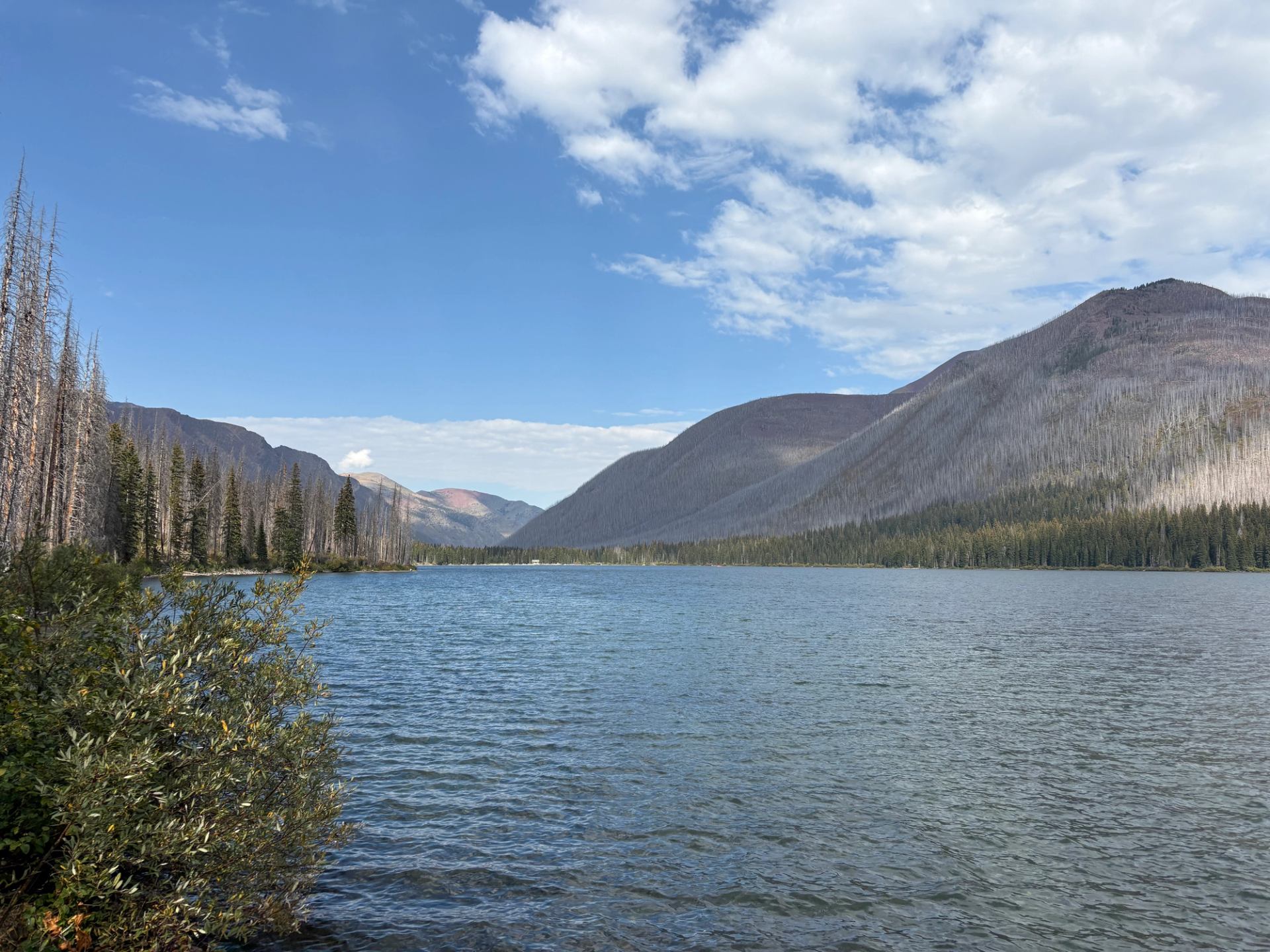 Mountain lake with calm water, forested shoreline and distant hills under partly cloudy sky.