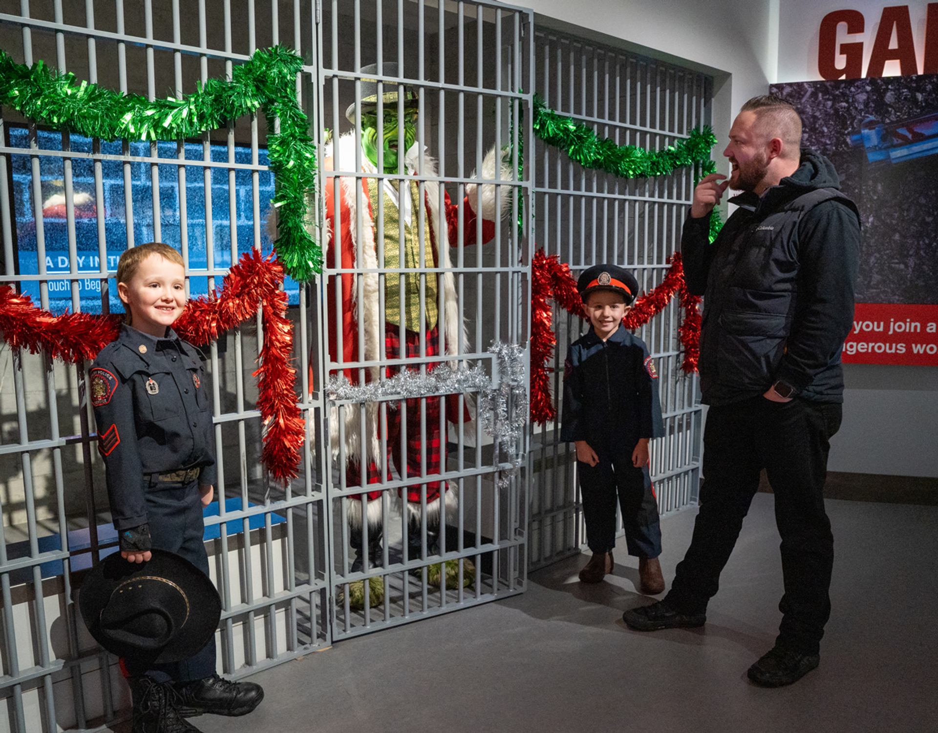 Costumed Grinch stands behind decorated jail bars beside two children and an adult.